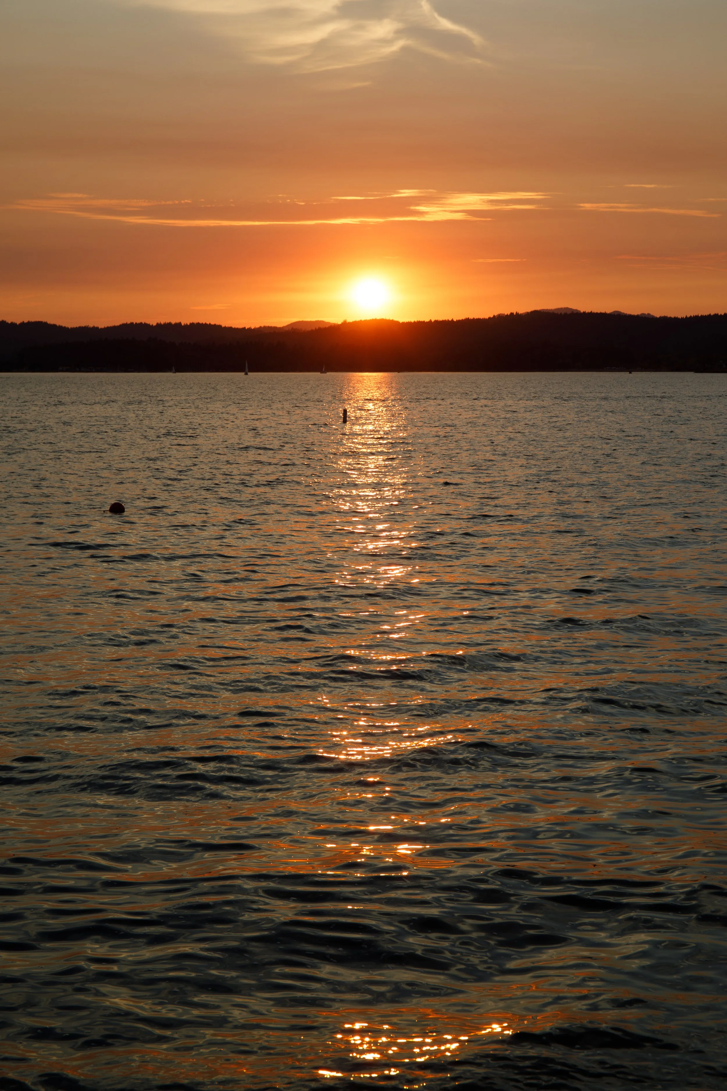 Sunset over the water with reflections and small sailboats in the distance.