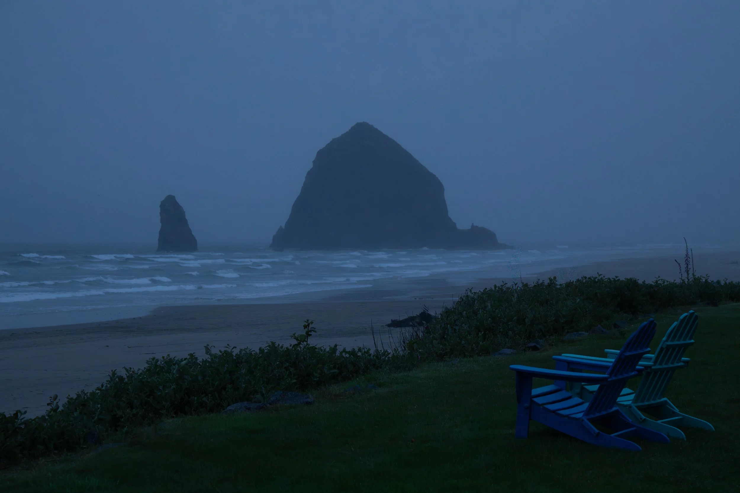 A foggy beach scene at dusk with two large rocks emerging from the ocean, and a grassy area with three empty Adirondack chairs facing the water.