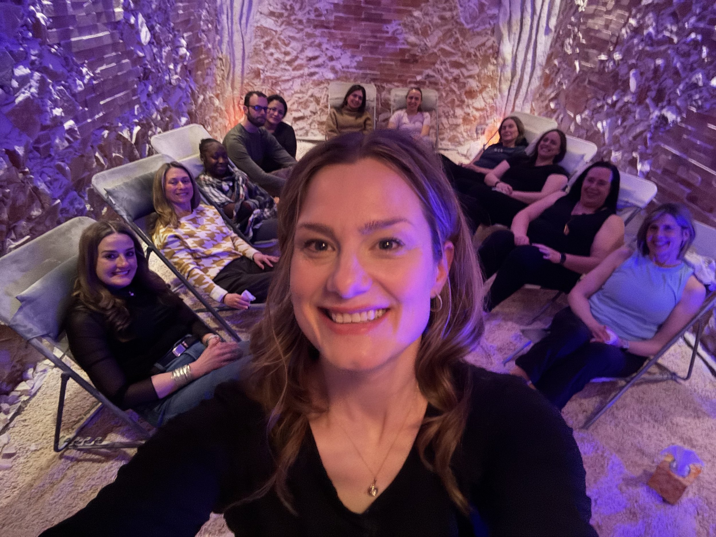 A group of people lounging on chairs in a room with brick walls and purple ambient lighting, with one person taking a selfie in the foreground.