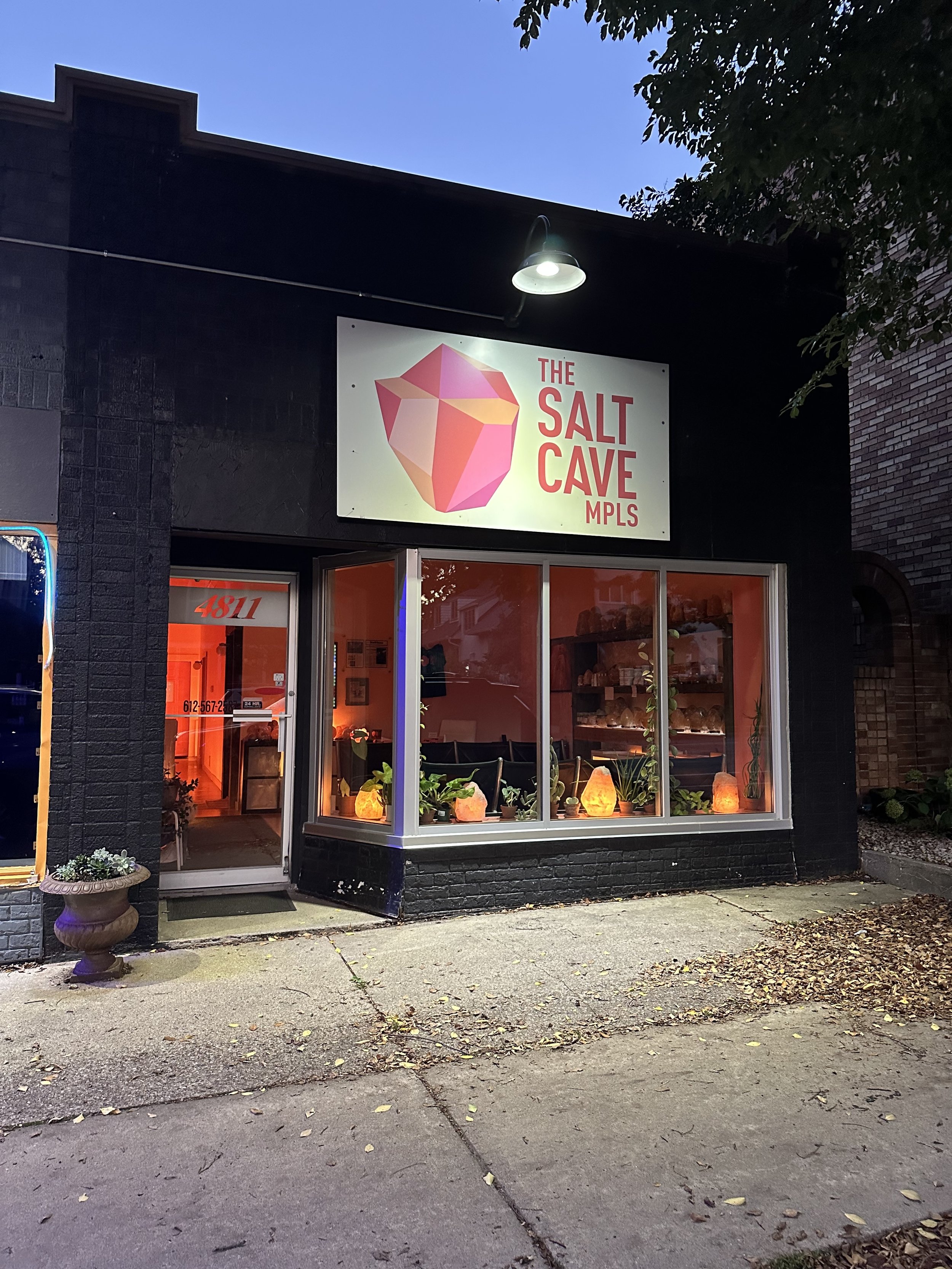 Storefront with a sign reading 'The Salt Cave MPLS' featuring a geometric salt crystal logo, located in a black brick building, illuminated from above, with a display window showing salt lamps and plants, and a sidewalk with fallen leaves in front.