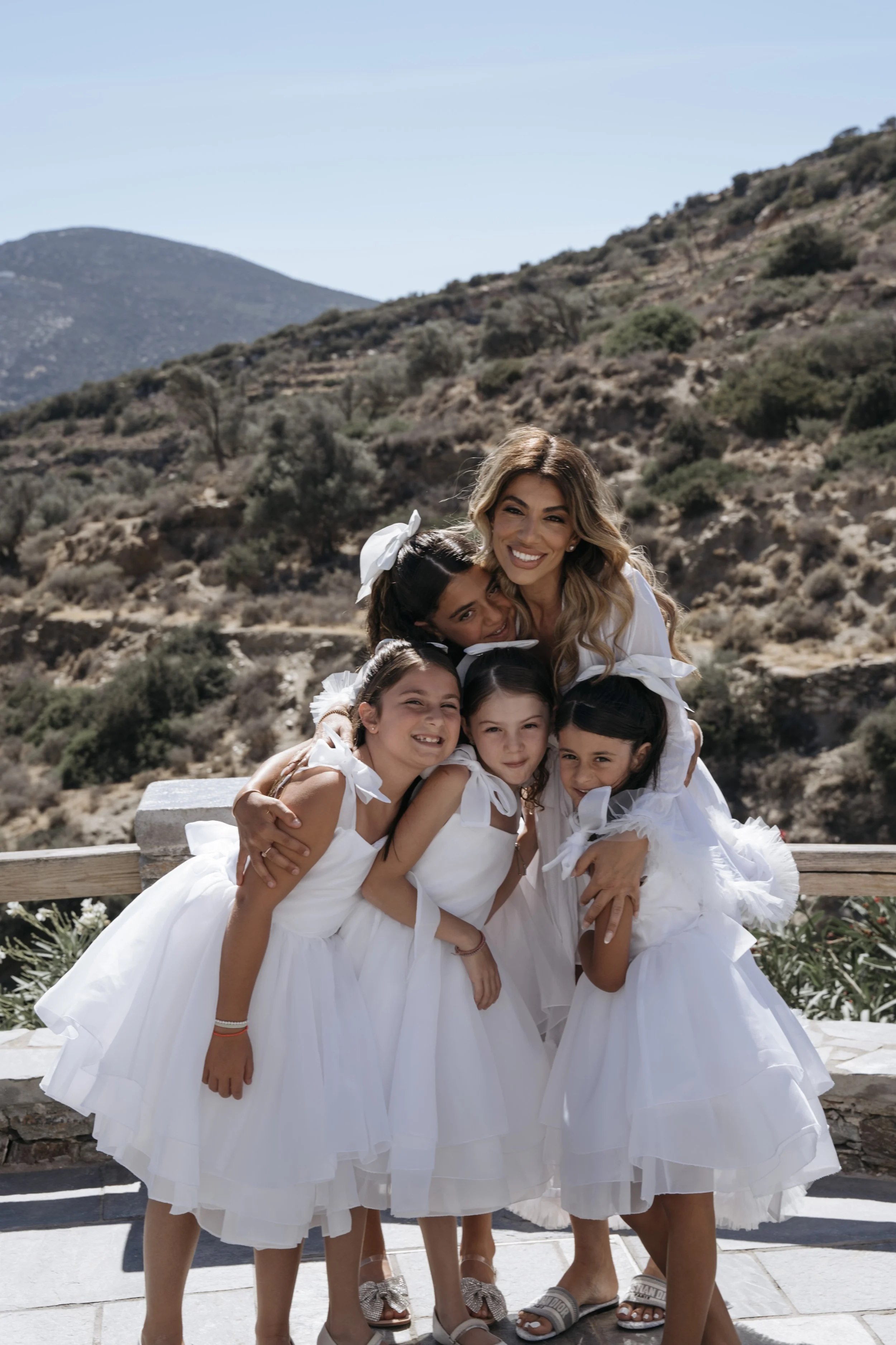 Bride during mid-morning bridal prep in her robe with dressed flower girls, wearing soft, natural bridal makeup in Sifnos, Greece.