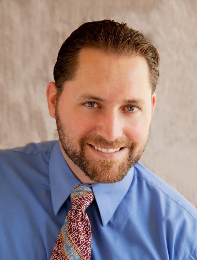 Portrait of a man with brown hair and a beard, smiling, wearing a blue dress shirt and patterned tie, against a light background.