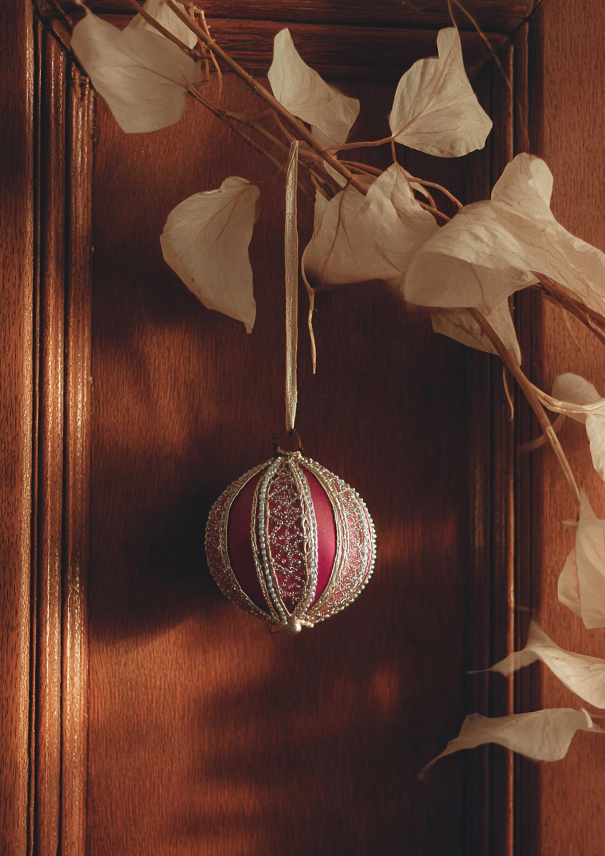 Decorative red Christmas ornament with gold detailing hanging from a branch of dry, light-colored leaves against a wooden background.