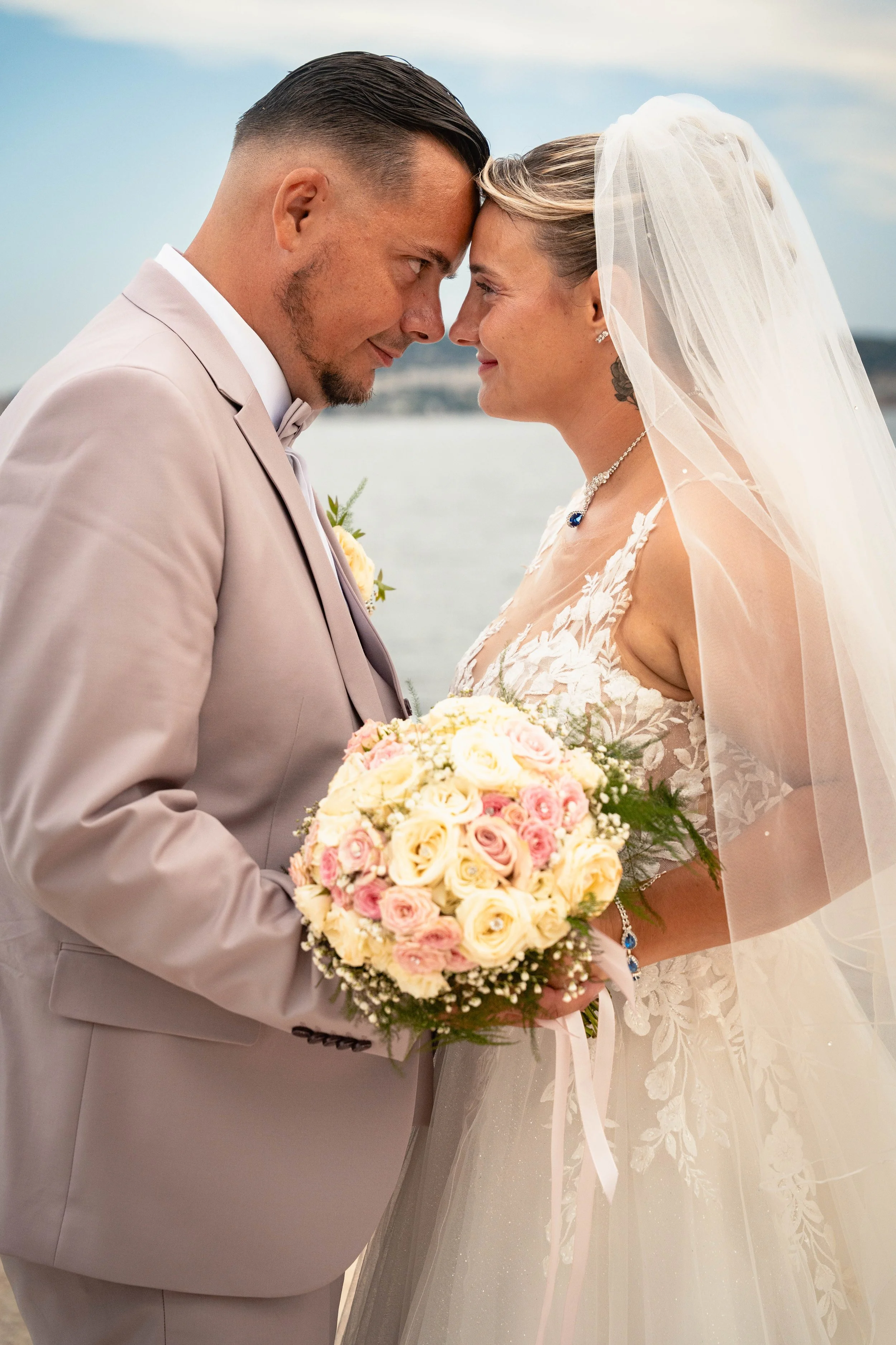 Un couple en mariage échangeant un regard tendre, la femme porte une robe de mariée et un voile, lui porte un costume clair, ils tiennent un bouquet de fleurs roses, avec un fond de ciel et d'eau.