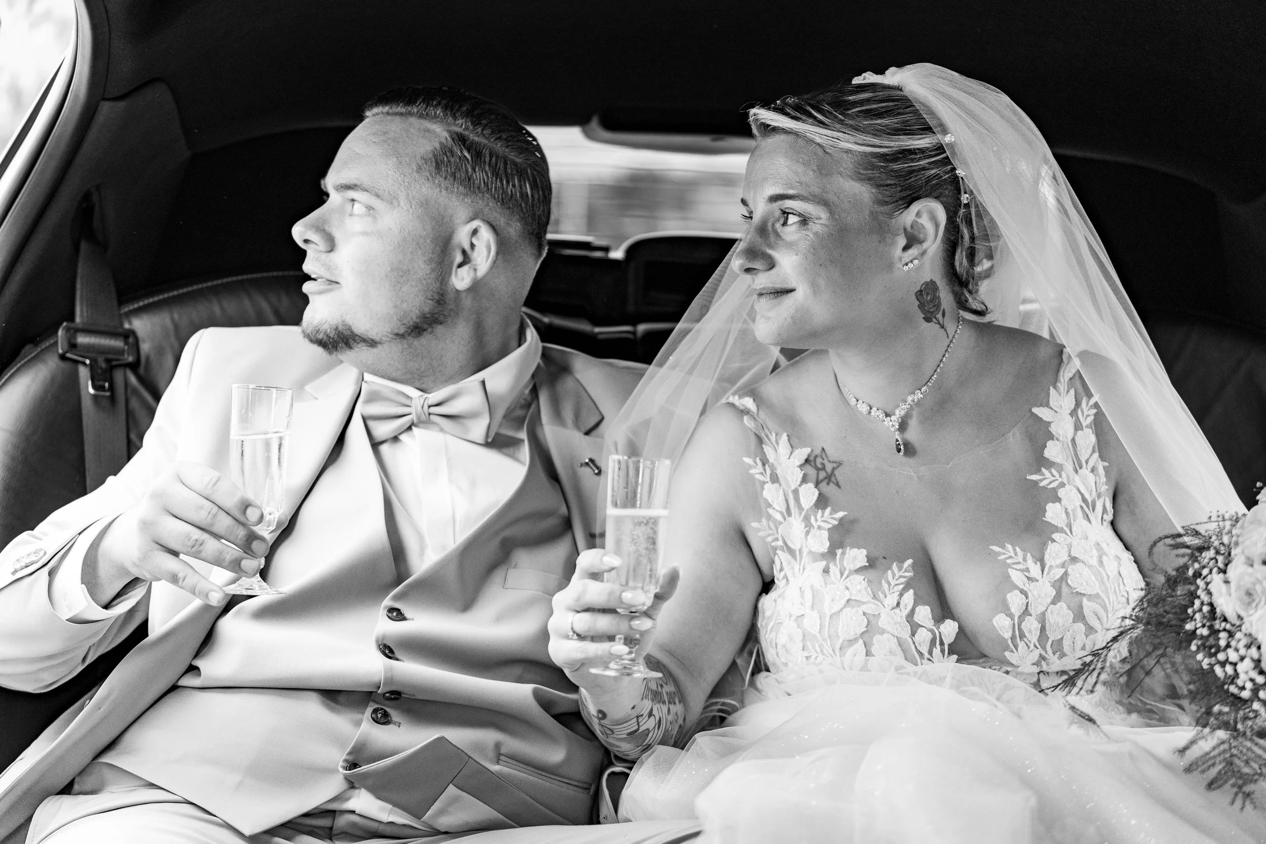 Un couple de mariés assis dans une voiture, la femme porte une robe de mariée avec une voilette, et tient un verre de champagne. L'homme porte un costume avec nœud papillon, tous deux sourient et regardent vers l'extérieur.