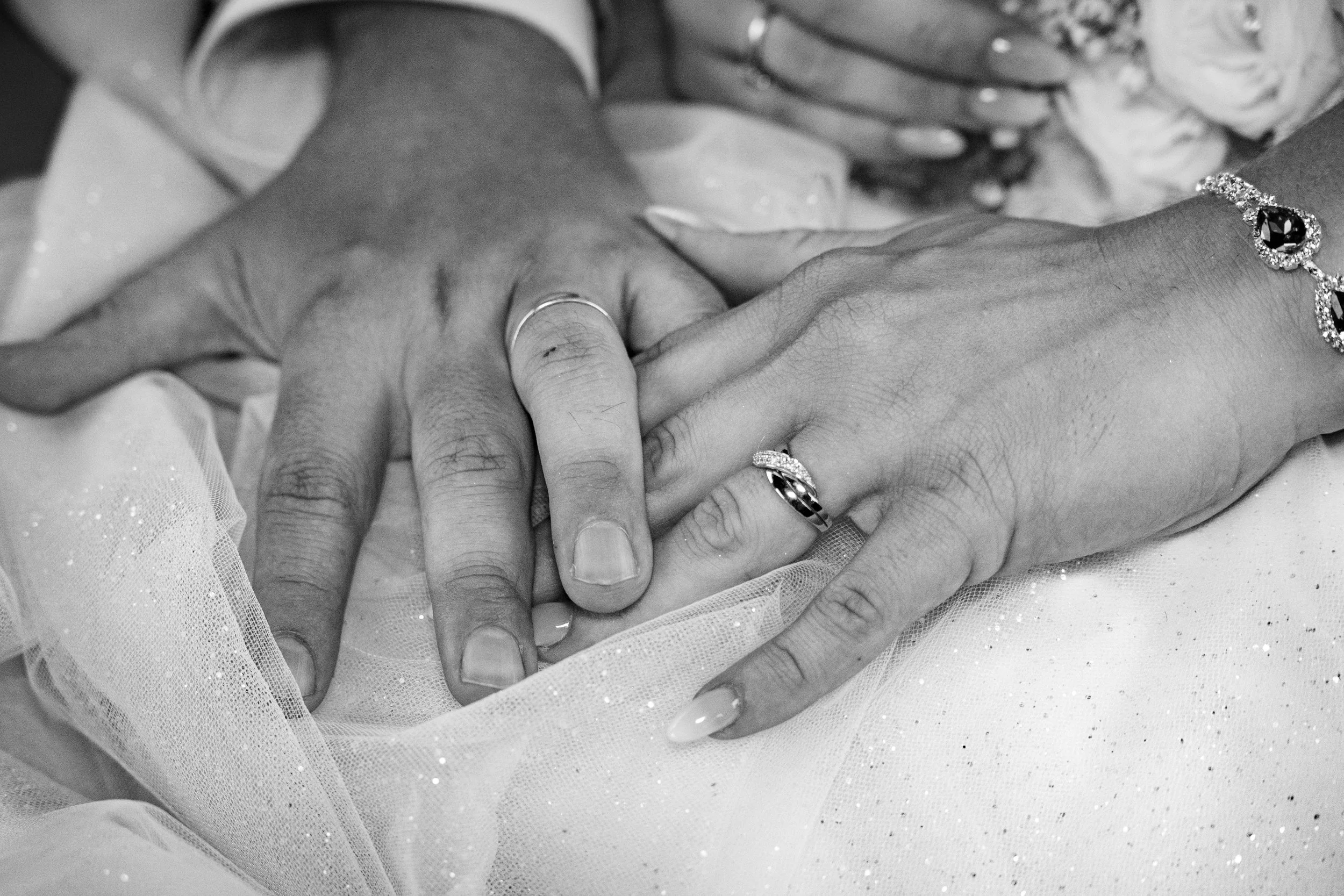Gros plan en noir et blanc de deux mains entrelacées, portant des alliances, avec des épaules et des bras visibles en arrière-plan. L'une porte une bague avec un grand pendentif noir entouré de pierres précieuses.