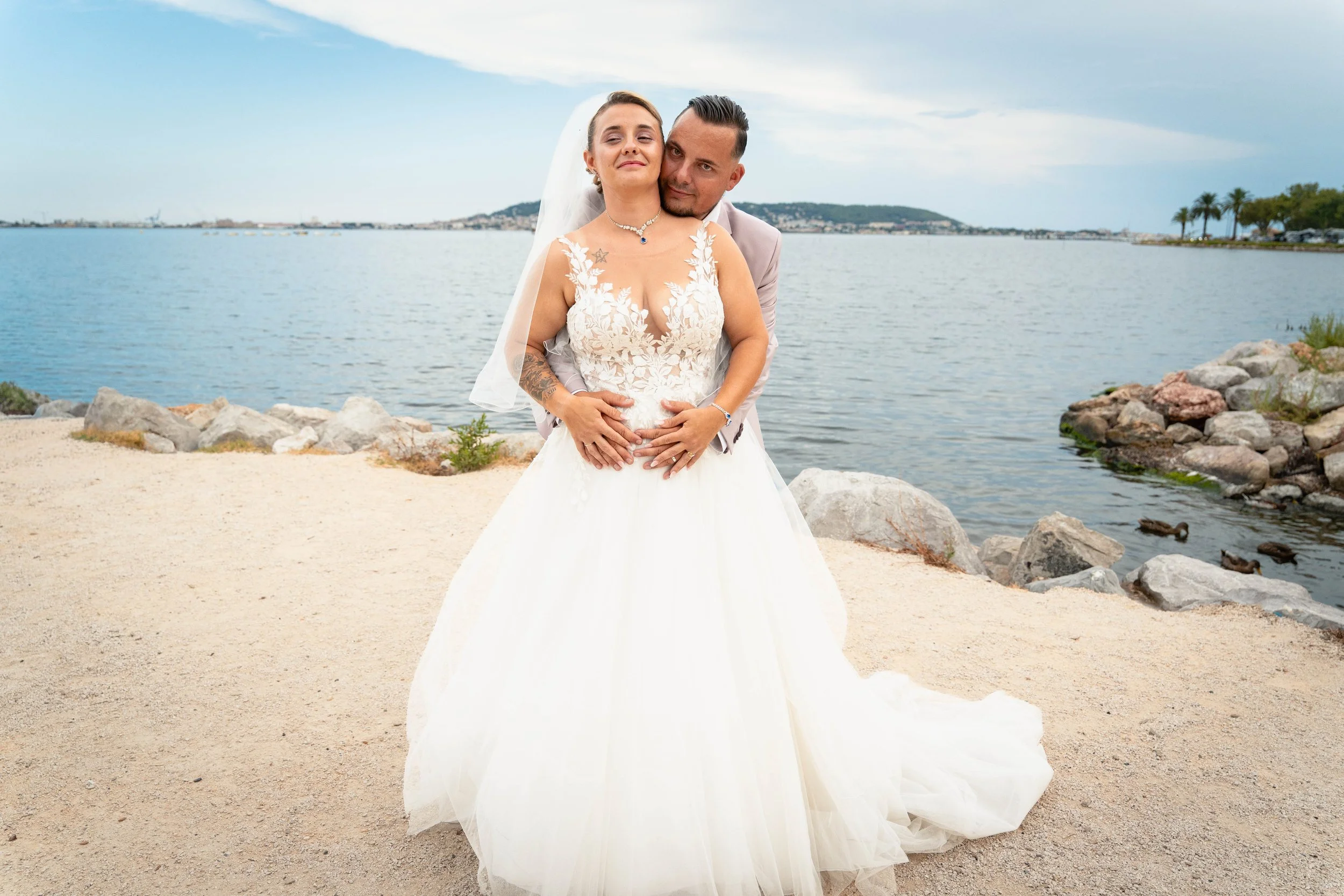 Un couple en mariage pose au bord de l'étang de thau avec Sète et un ciel nuageux en arrière-plan.