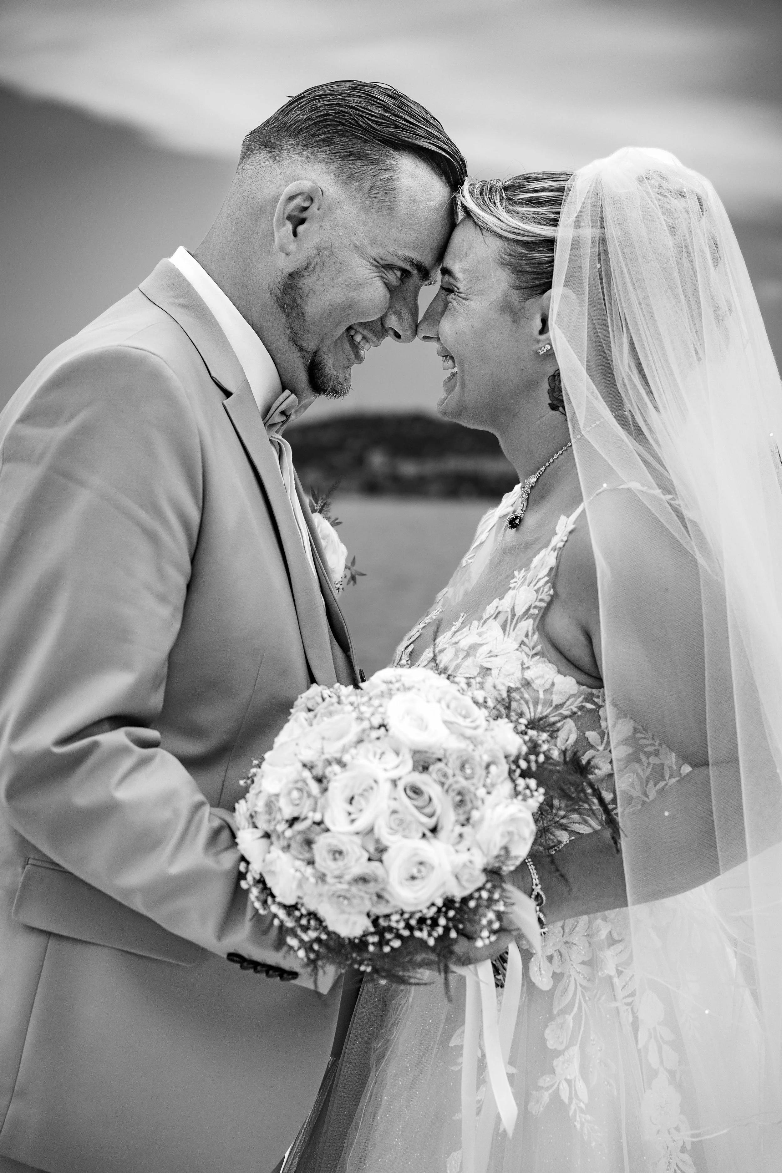 Un couple de mariés souriants se touche le front lors de leur mariage en plein air, en noir et blanc.