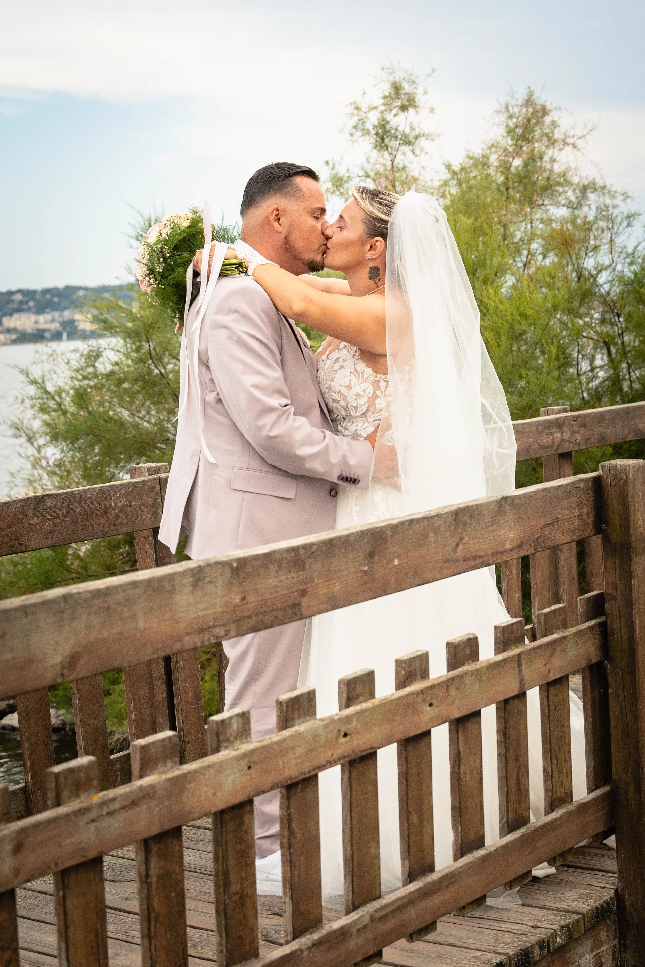 Un couple en mariage s'embrassant sur un pont en bois, avec des arbres et un ciel nuageux en arrière-plan.