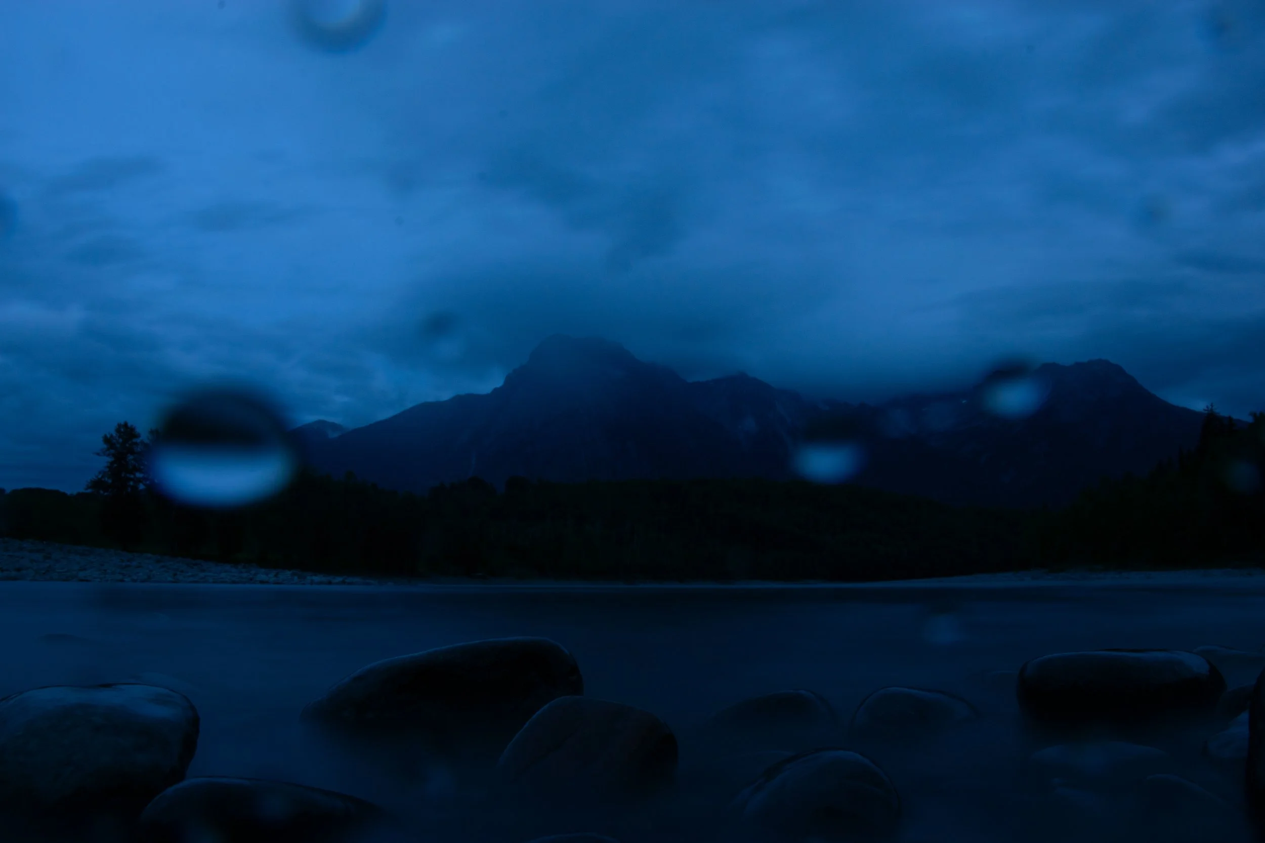 Dark, cloudy sky over a mountain range, with raindrops on a window in the foreground and rocks near a body of water.