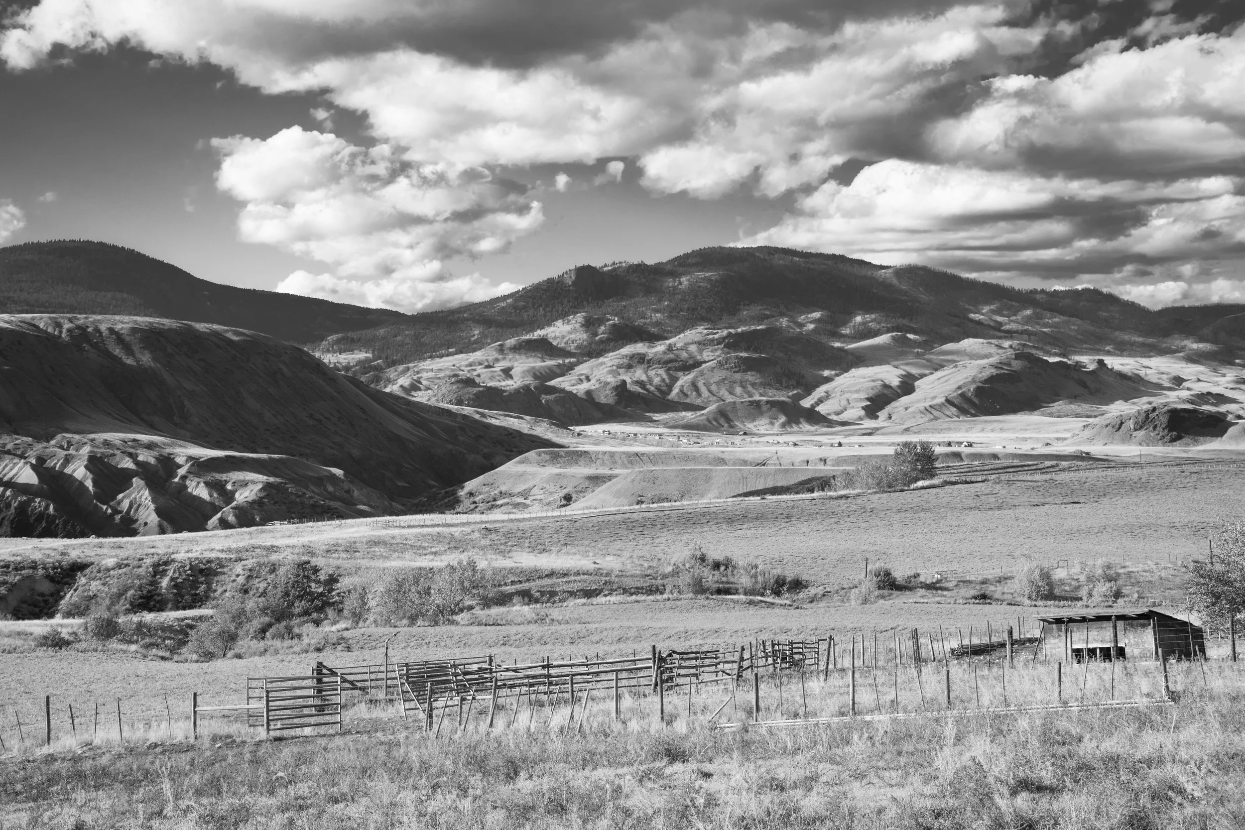 A panoramic view of rolling hills and mountains under a partly cloudy sky, with fenced fields in the foreground.