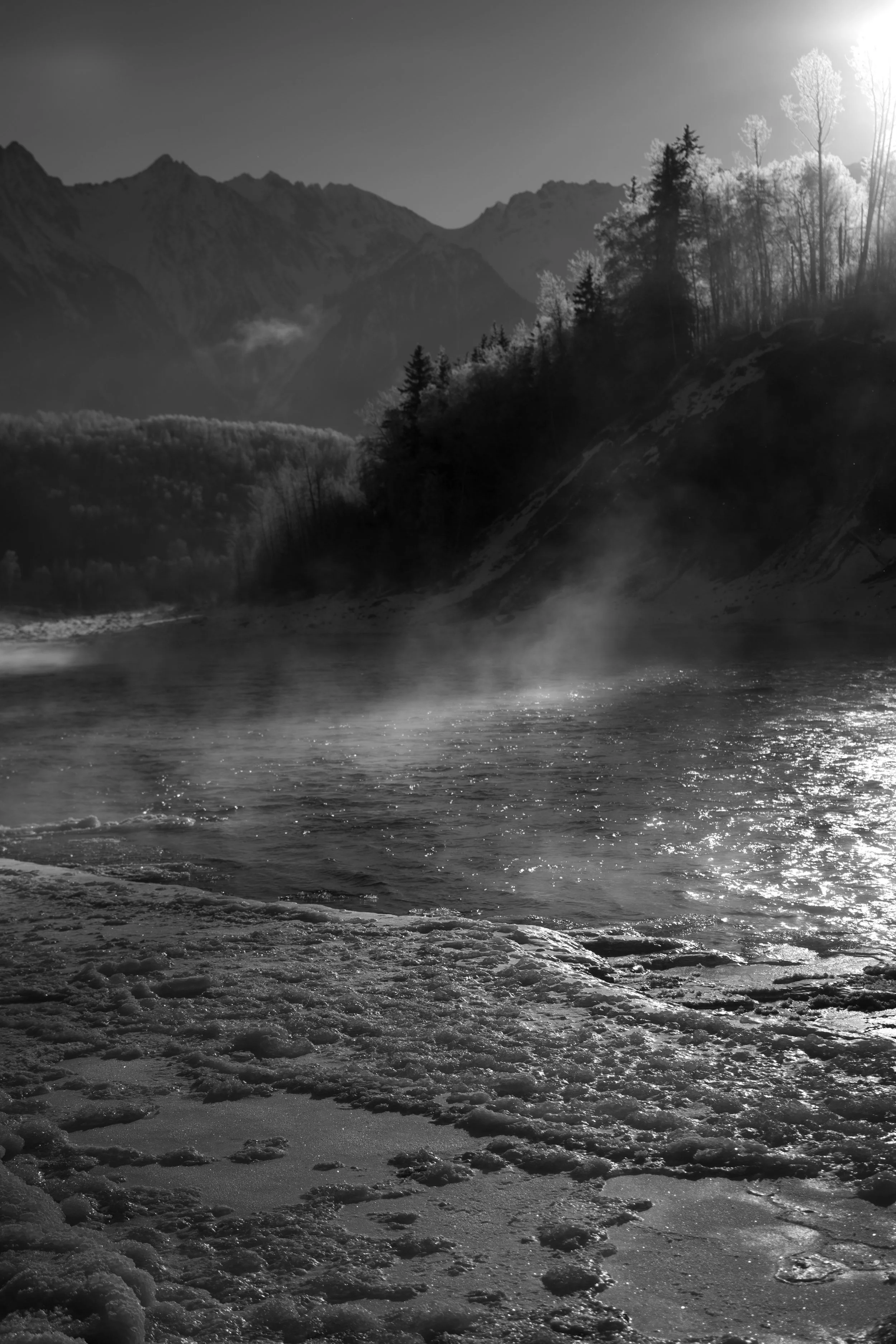 Black and white photo of a mountain landscape with a river in the foreground, mist rising from the water, and trees on the hillside, with the sun shining from the upper right corner.