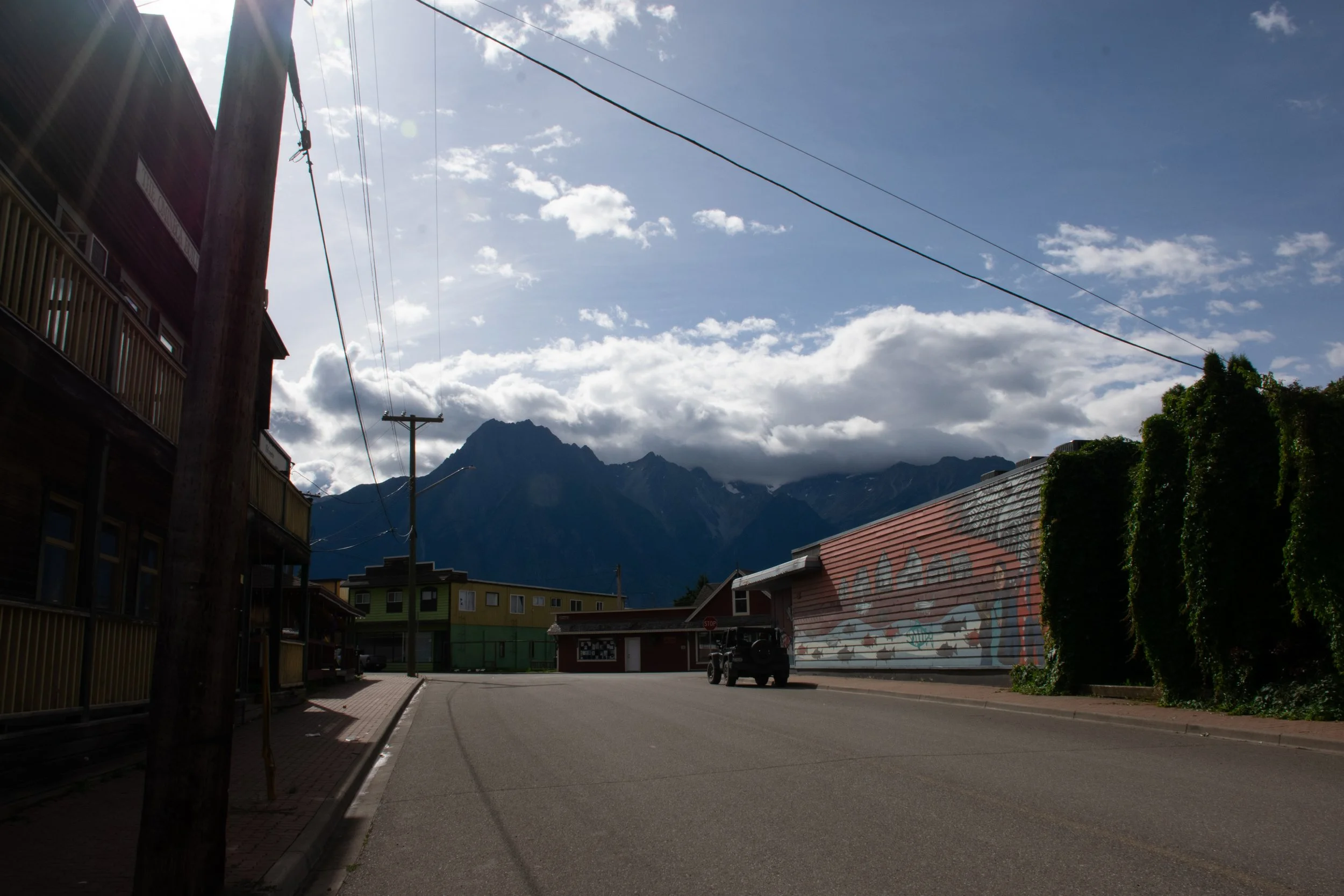 Street view of a small town with mountains in the background, a building with murals on the right, power lines running overhead, and a wooden utility pole on the left. Cloudy sky with patches of blue.