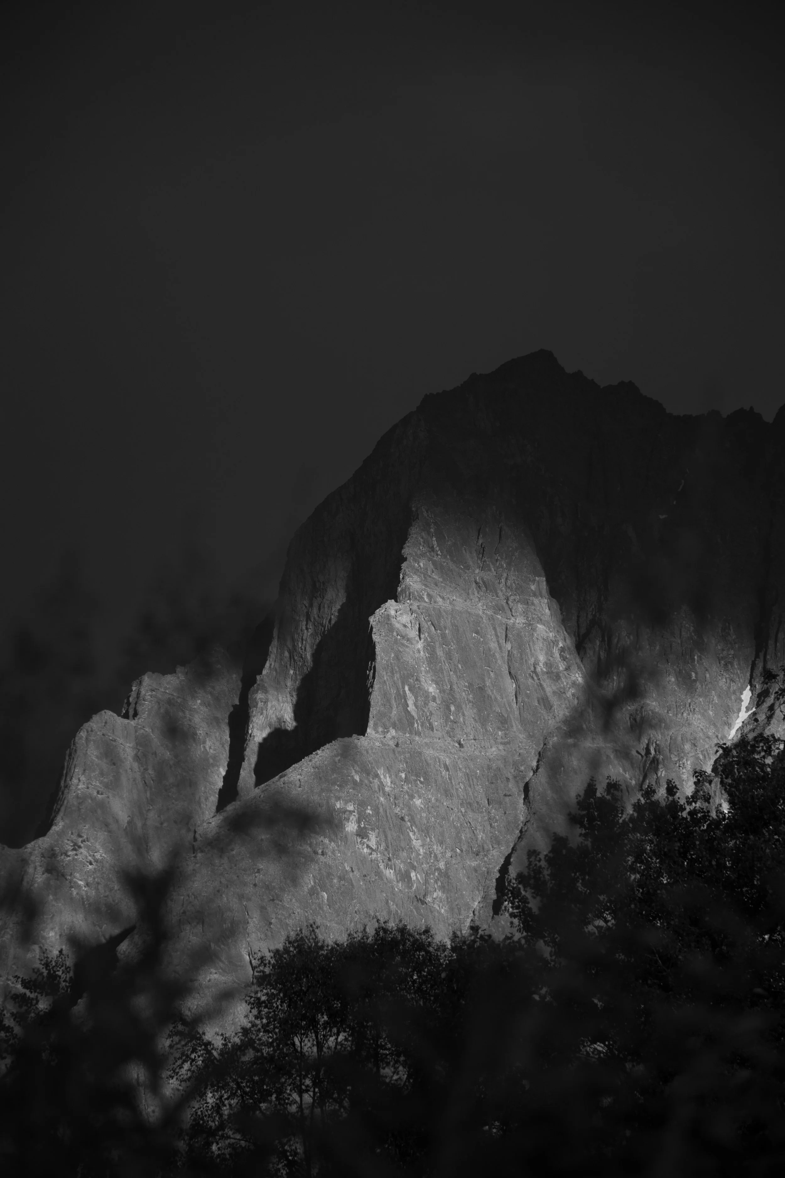 A black and white image of rugged mountain peaks with trees at the base, illuminated with a light source highlighting the rocks and cliffs.