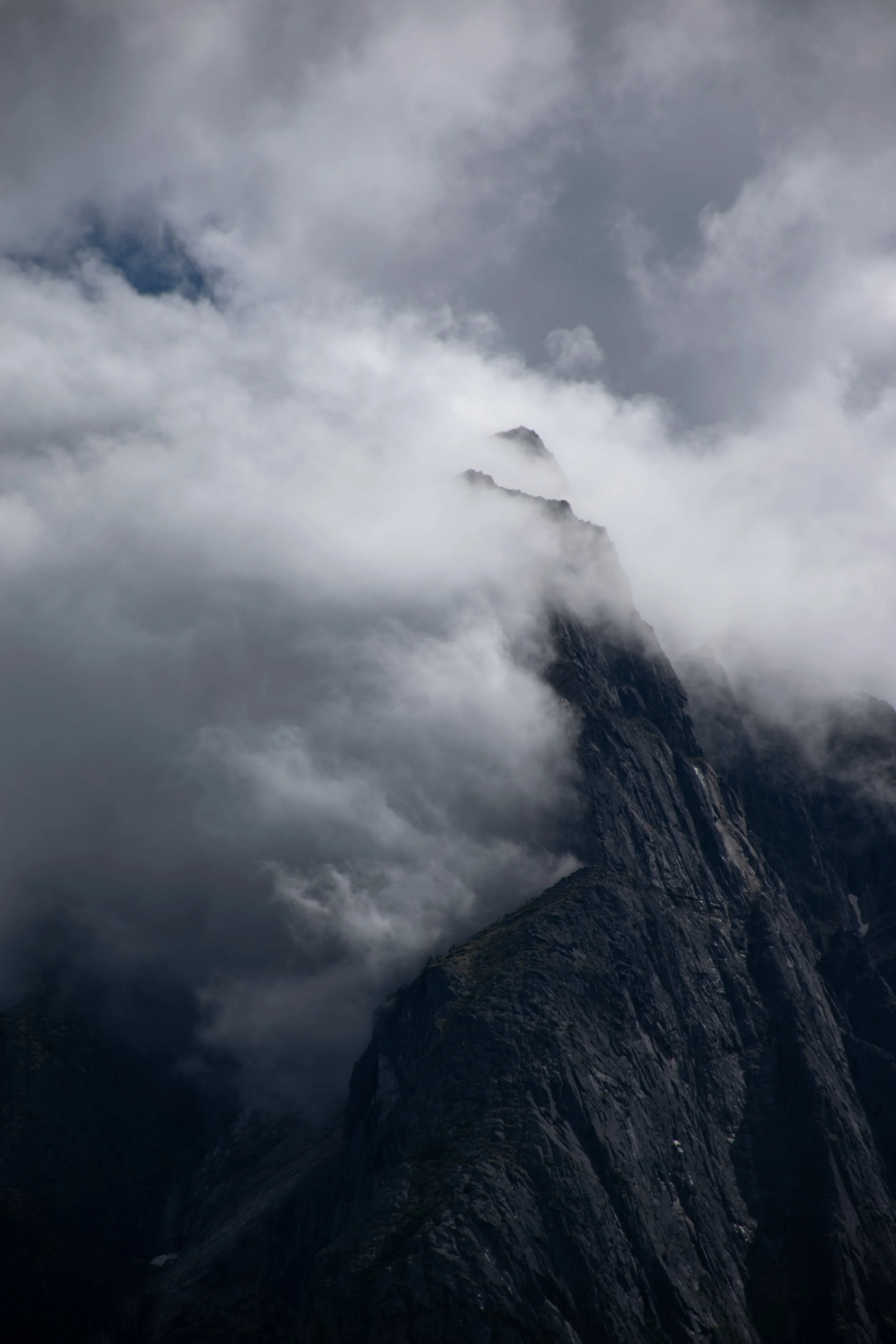 A steep mountain peak partially covered in clouds and fog, with rugged, rocky terrain.