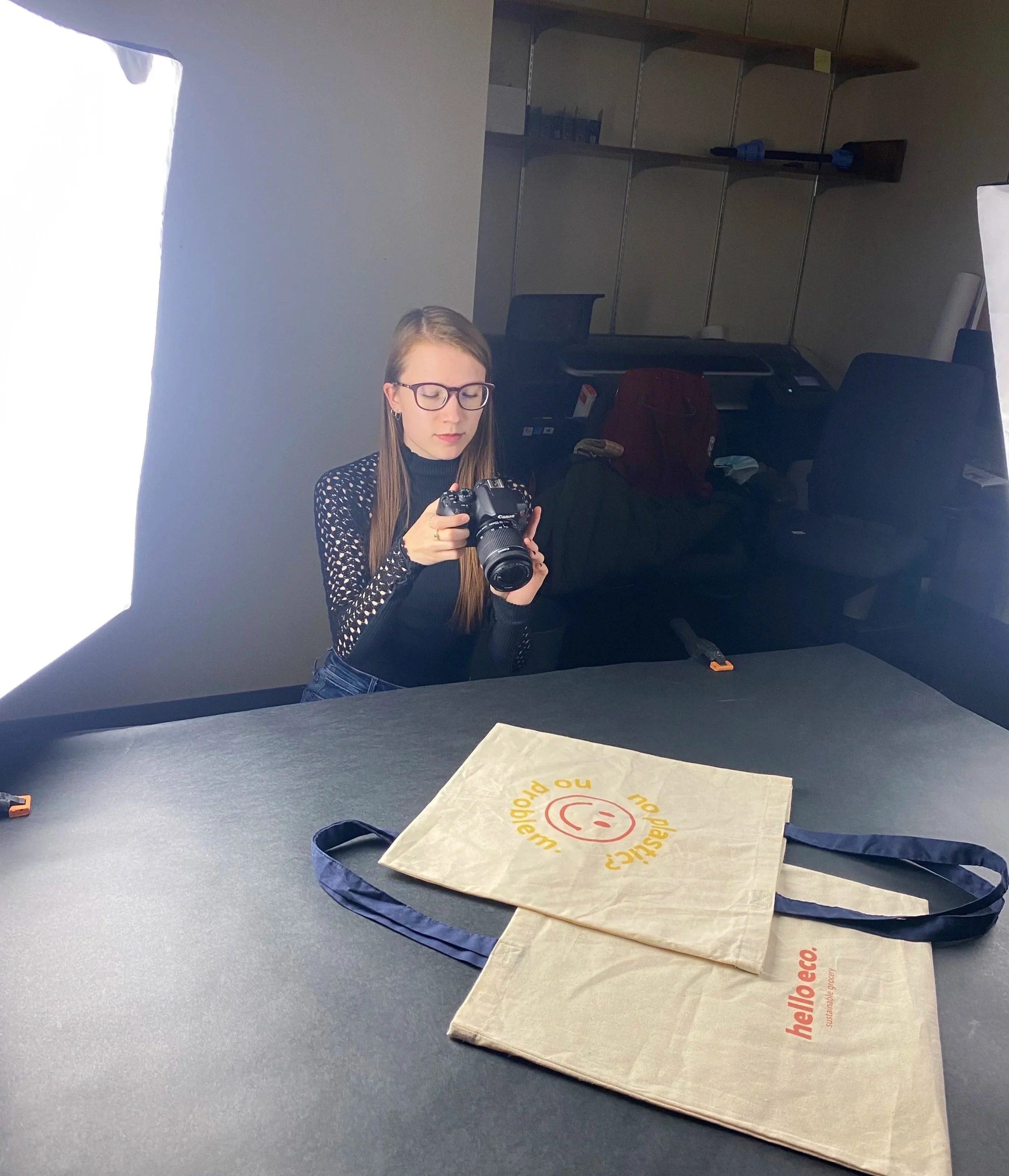 A woman wearing glasses and a black shirt captures a photo with her camera. A light backlights the scene, and in the background, there are shelves with various items.