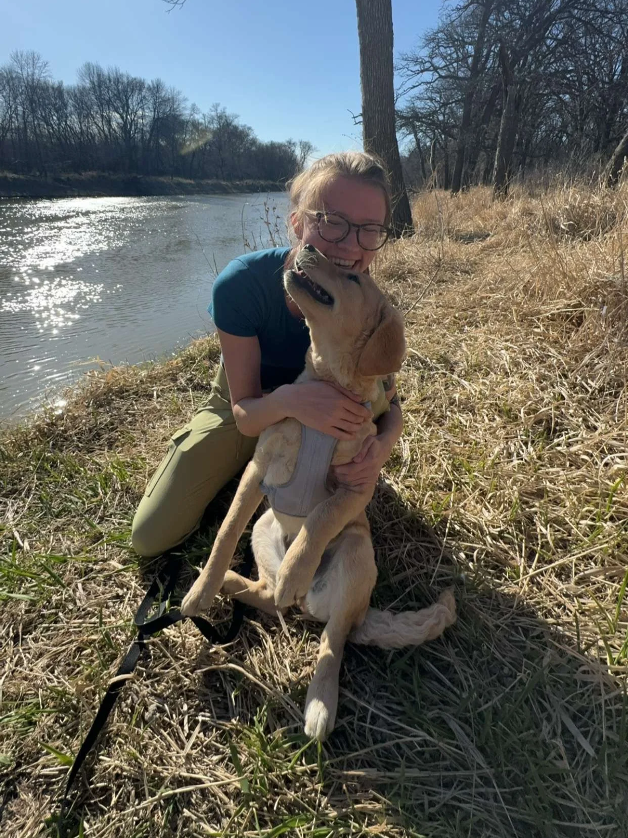 A woman sitting on dry grass beside a river, hugging a large yellow Labrador Retriever dog. The woman is wearing glasses, a blue shirt, and khaki pants, smiling as the dog looks happy. The background shows leafless trees and a bright blue sky.