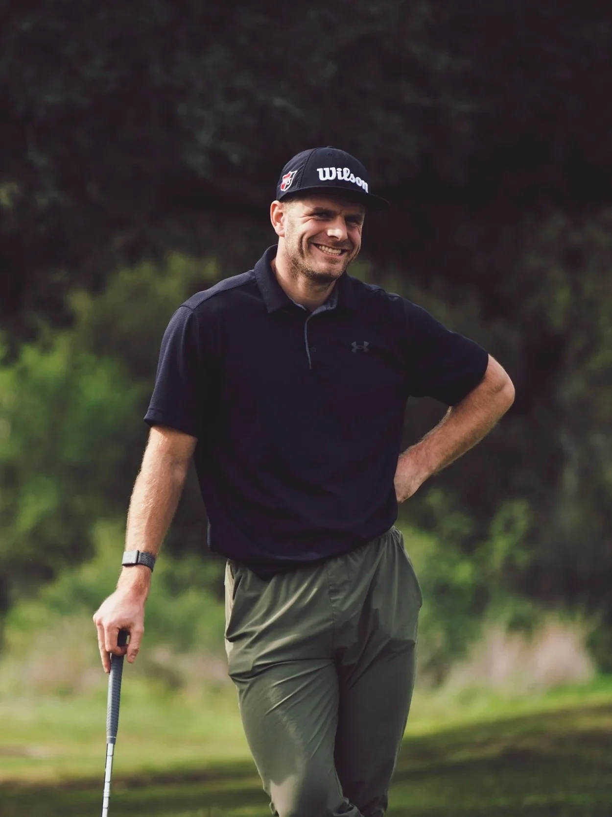 A man smiling while playing golf outdoors. He is holding a golf club, wearing a black cap with 'Wilson' written on it, a black polo shirt, and khaki pants, with greenery in the background.