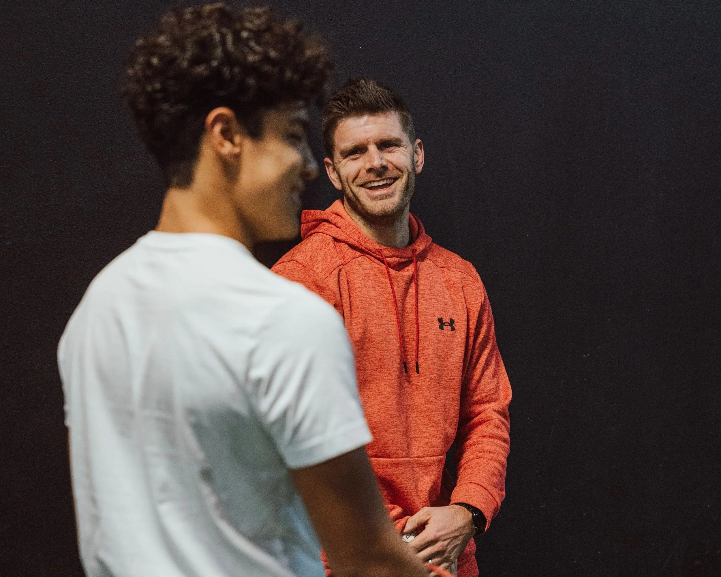 Two men are talking to each other, one with dark brown skin and curly hair, and another with light skin and short dark hair. The man with short dark hair is smiling and wearing an orange hoodie. The other man is wearing a white shirt. They are standing against a dark background.