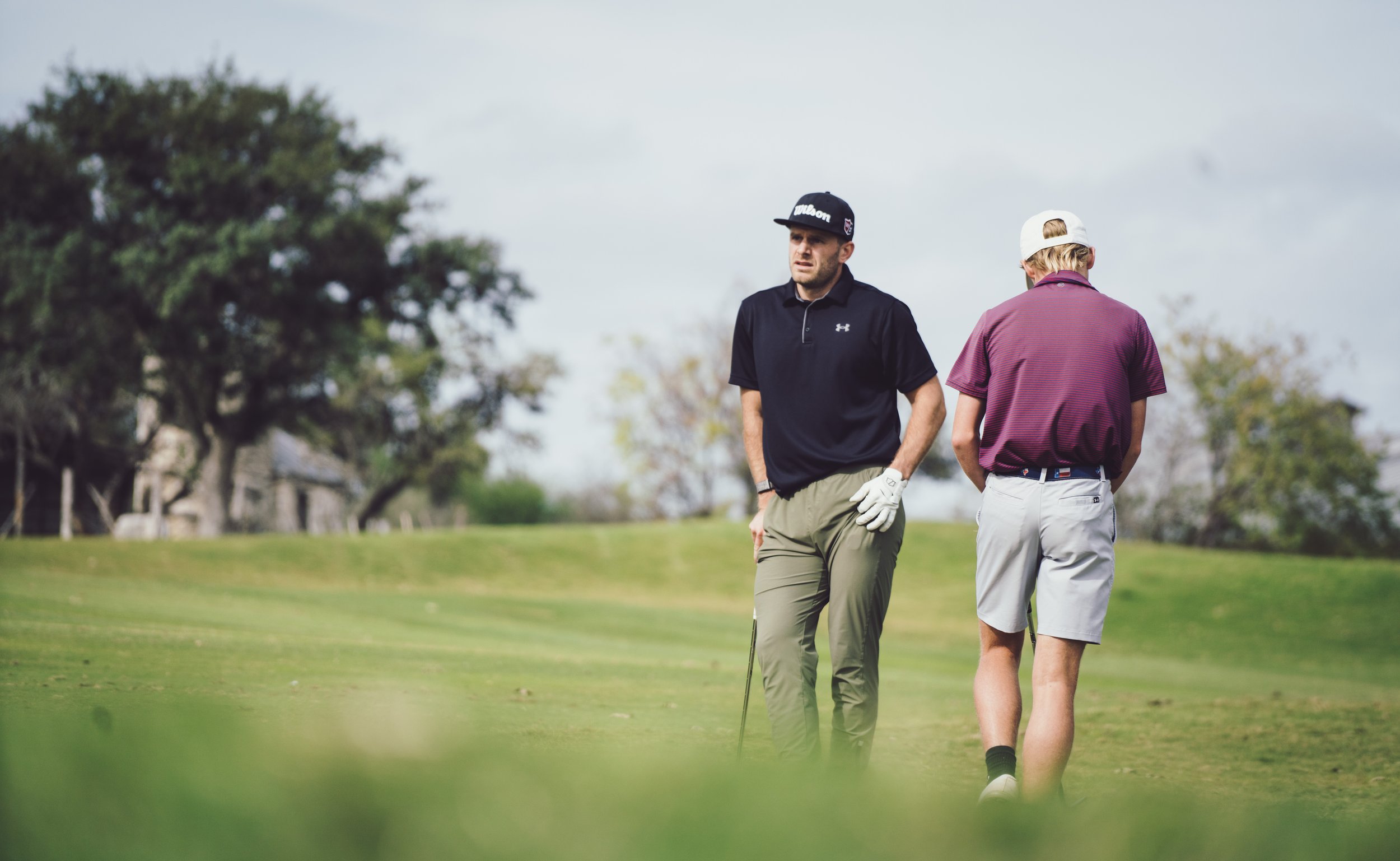 Two men walking on a golf course, one dressed in a black shirt and khaki pants, the other in a purple shirt and light gray shorts, with trees and cloudy sky in the background.