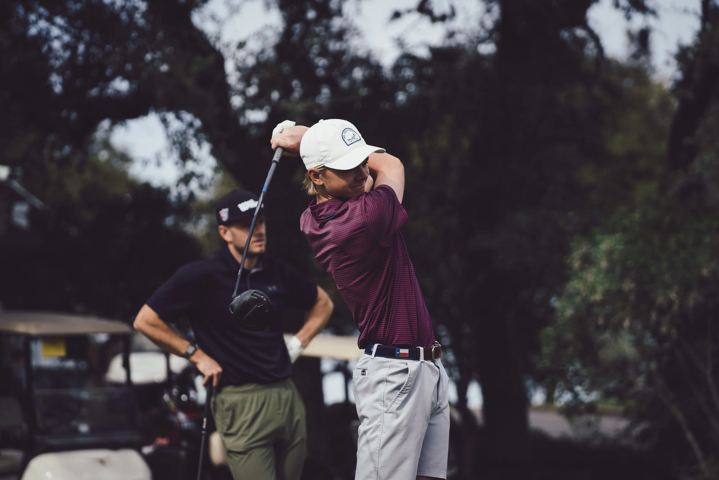 A woman in a striped shirt and white shorts is taking a swing with a golf club on a golf course, with a man in a black shirt and green pants standing behind her. There are trees in the background.
