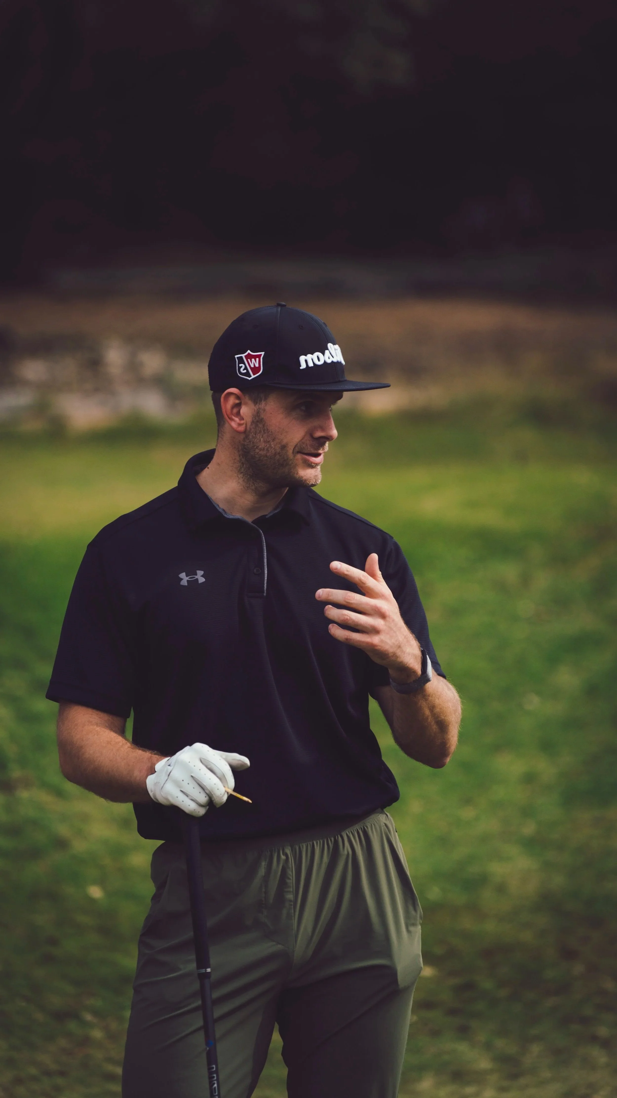 A man in a black golf cap, black Under Armour shirt, and khaki pants holding a golf club and a snack while talking outdoors on a cloudy day.