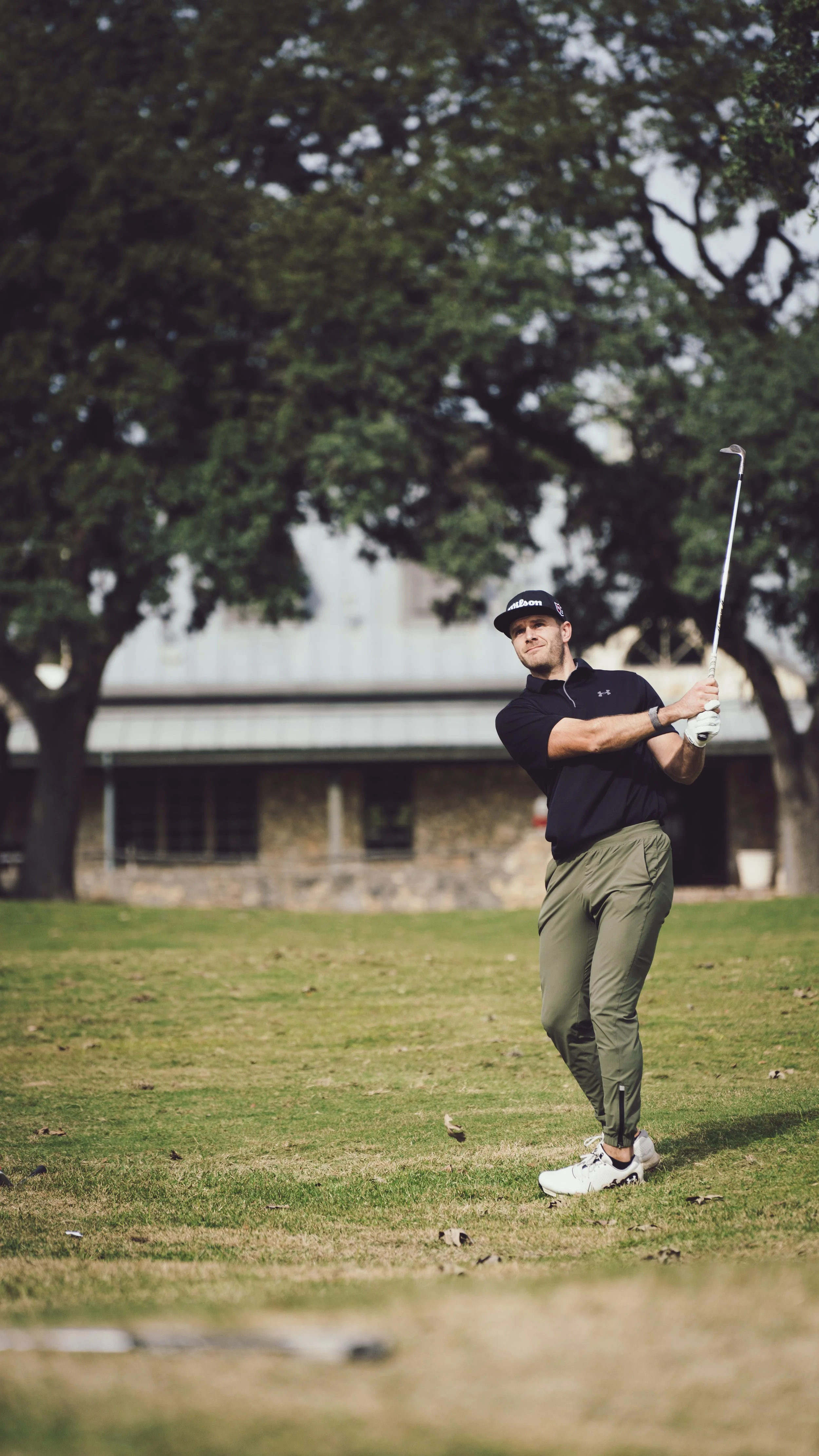 A man in golf attire swinging a golf club on a golf course with trees and a building in the background.