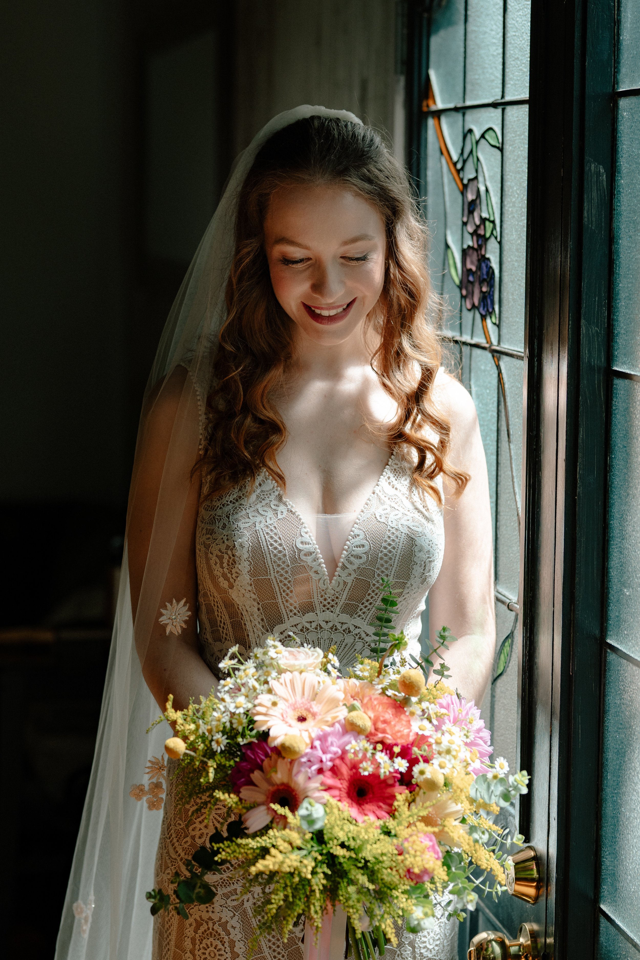 A bride with curly hair standing by a stained glass window, smiling and holding a large colorful bouquet of flowers.