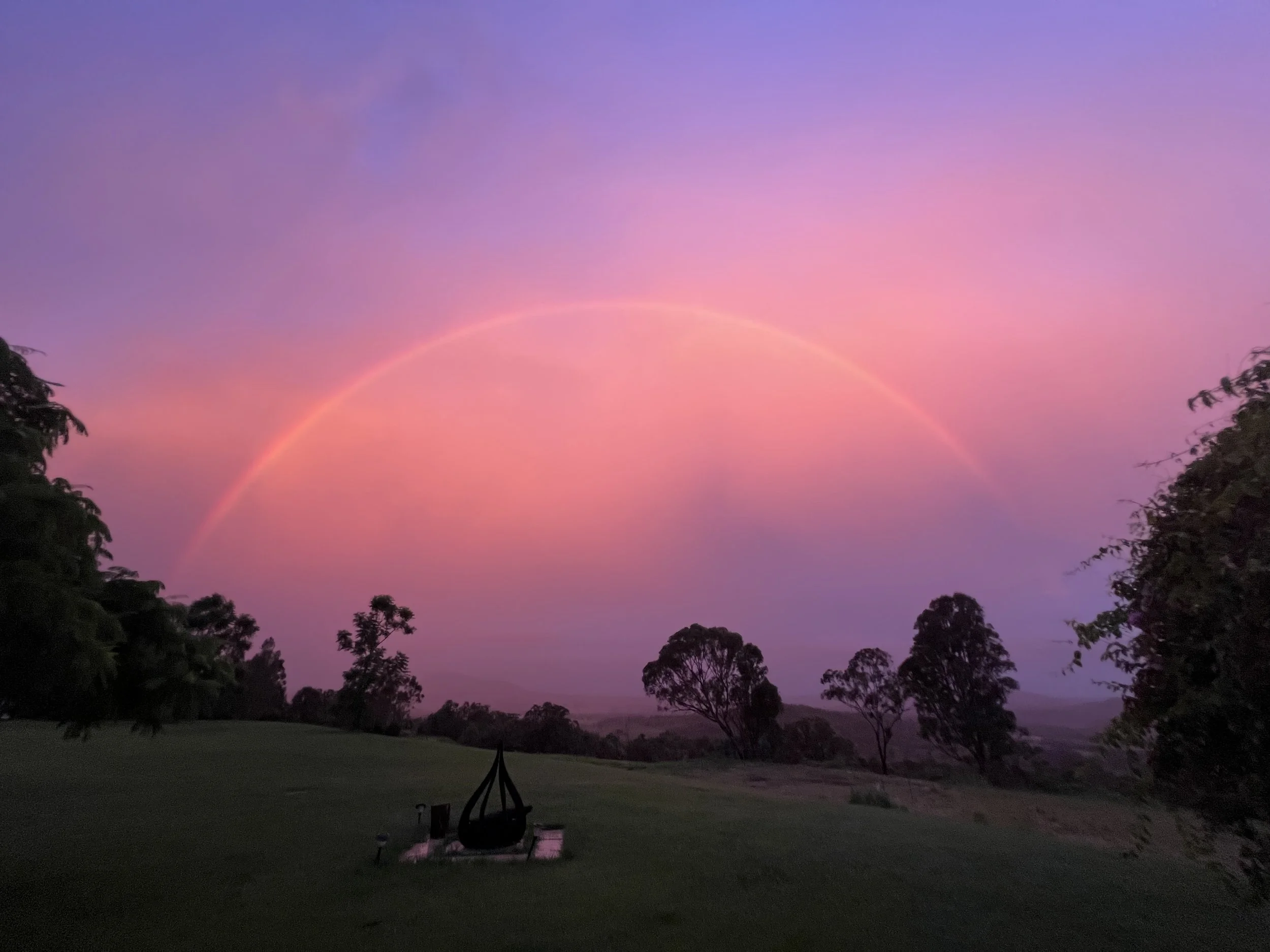 A colorful twilight sky featuring a rainbow and silhouettes of trees and a grassy field in the foreground.