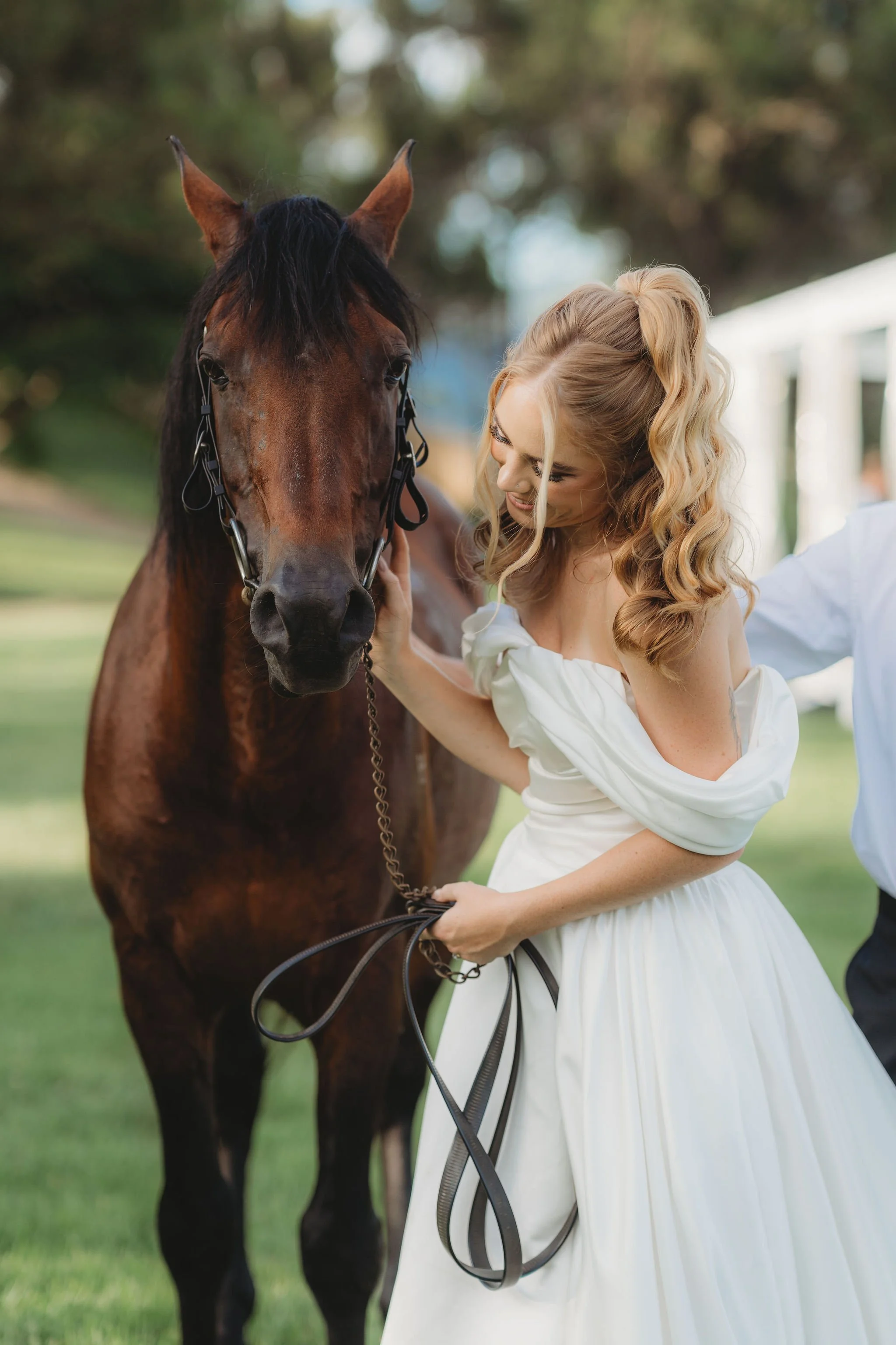 A woman in a white dress holding a brown horse's rein and smiling as she looks at the horse during daytime outdoors.