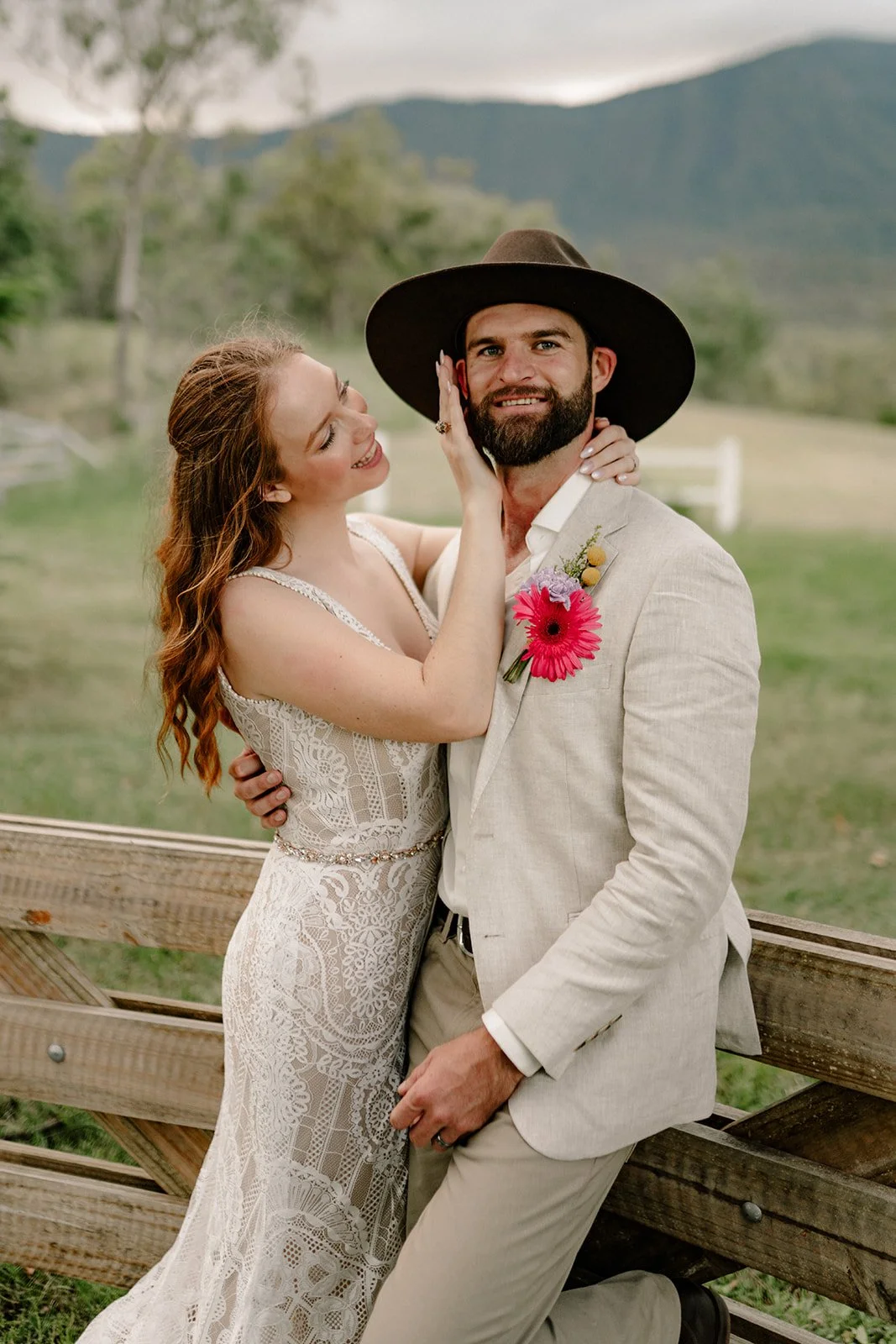 A woman in a lace wedding dress and a man in a light-colored suit with a black hat are embracing outdoors, with a scenic mountain background.