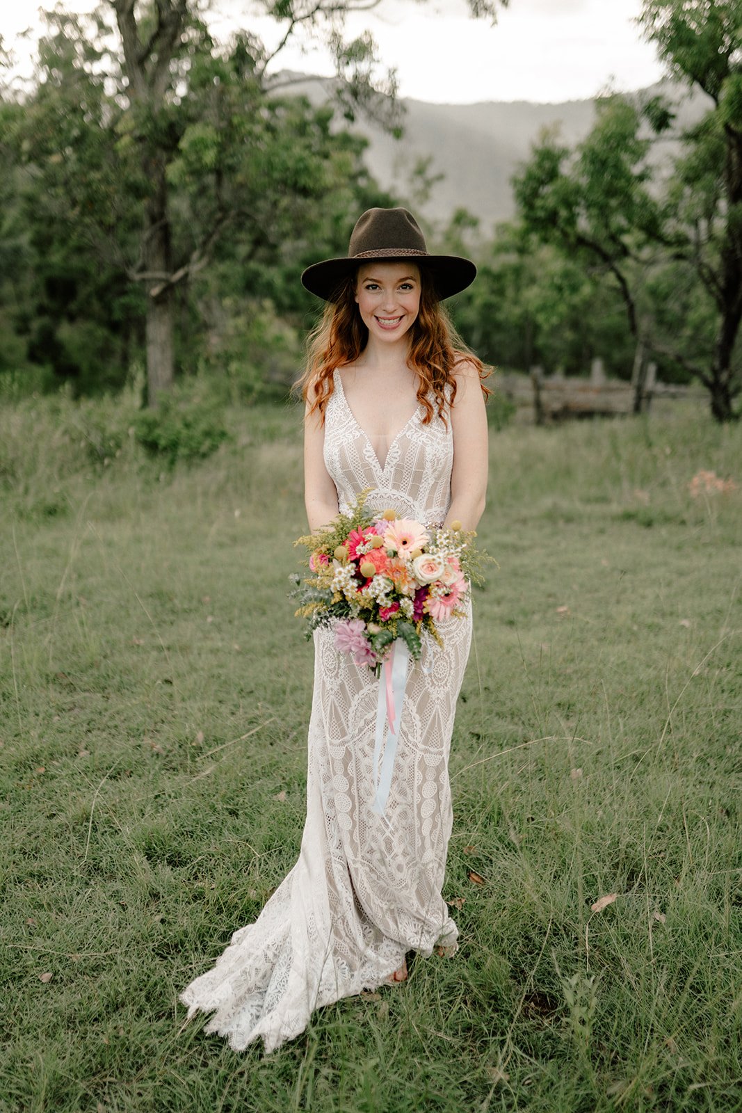A woman in a lace wedding dress with a deep neckline stands in a grassy field holding a colorful bouquet of flowers. She wears a wide-brimmed brown hat and has long, curly red hair. She is smiling and standing in front of trees with a mountain or hill in the background.