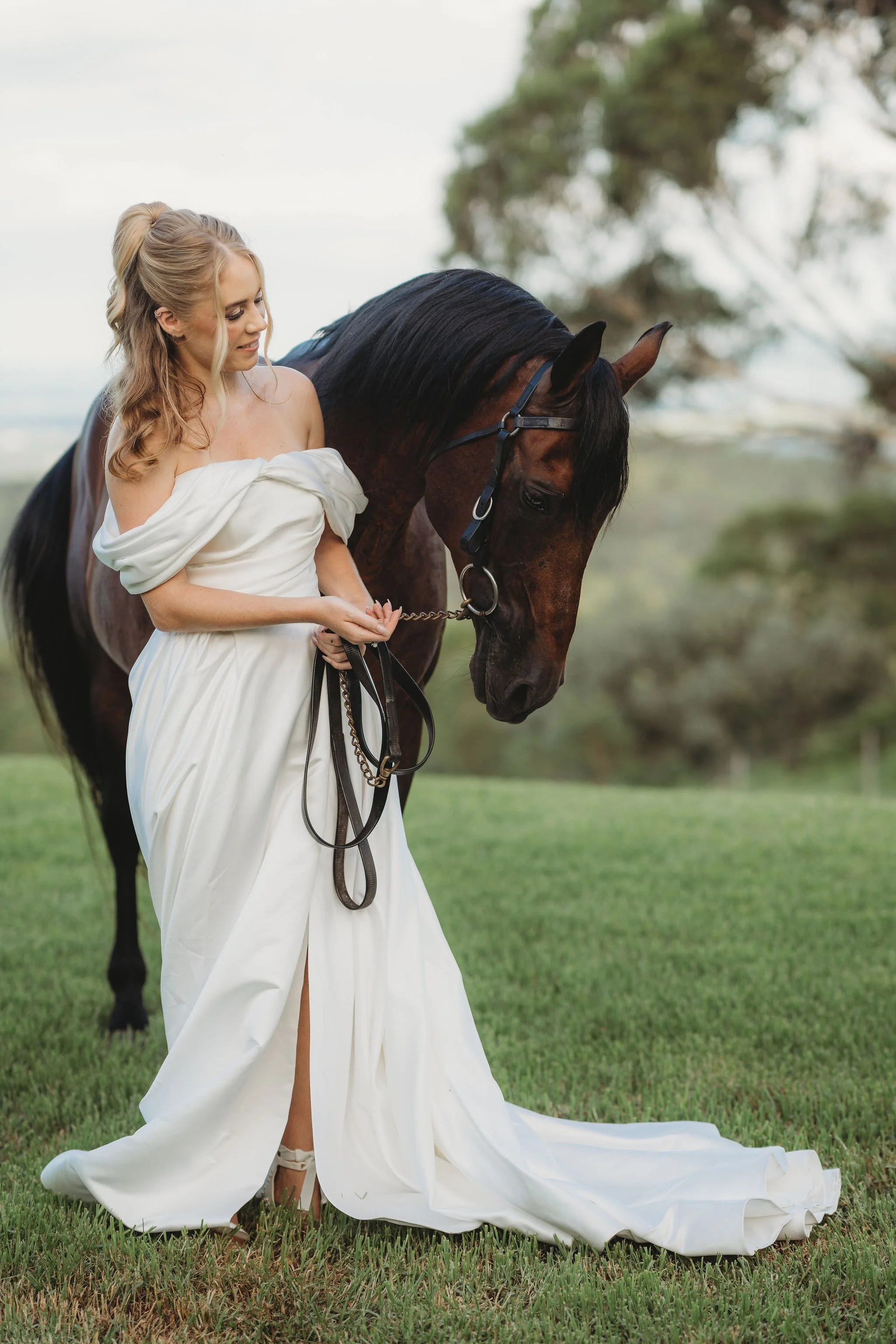 A woman with blonde hair in loose curls wearing a wedding dress holds a brown horse on a grassy field during daytime. The woman is smiling and looking down, and the horse's head is lowered, with trees in the background.