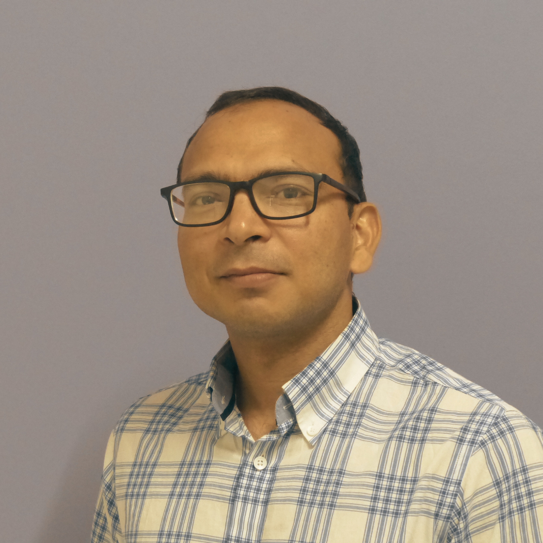An image of  man with short dark hair, glasses, wearing a blue and white checked shirt standing against a plain background