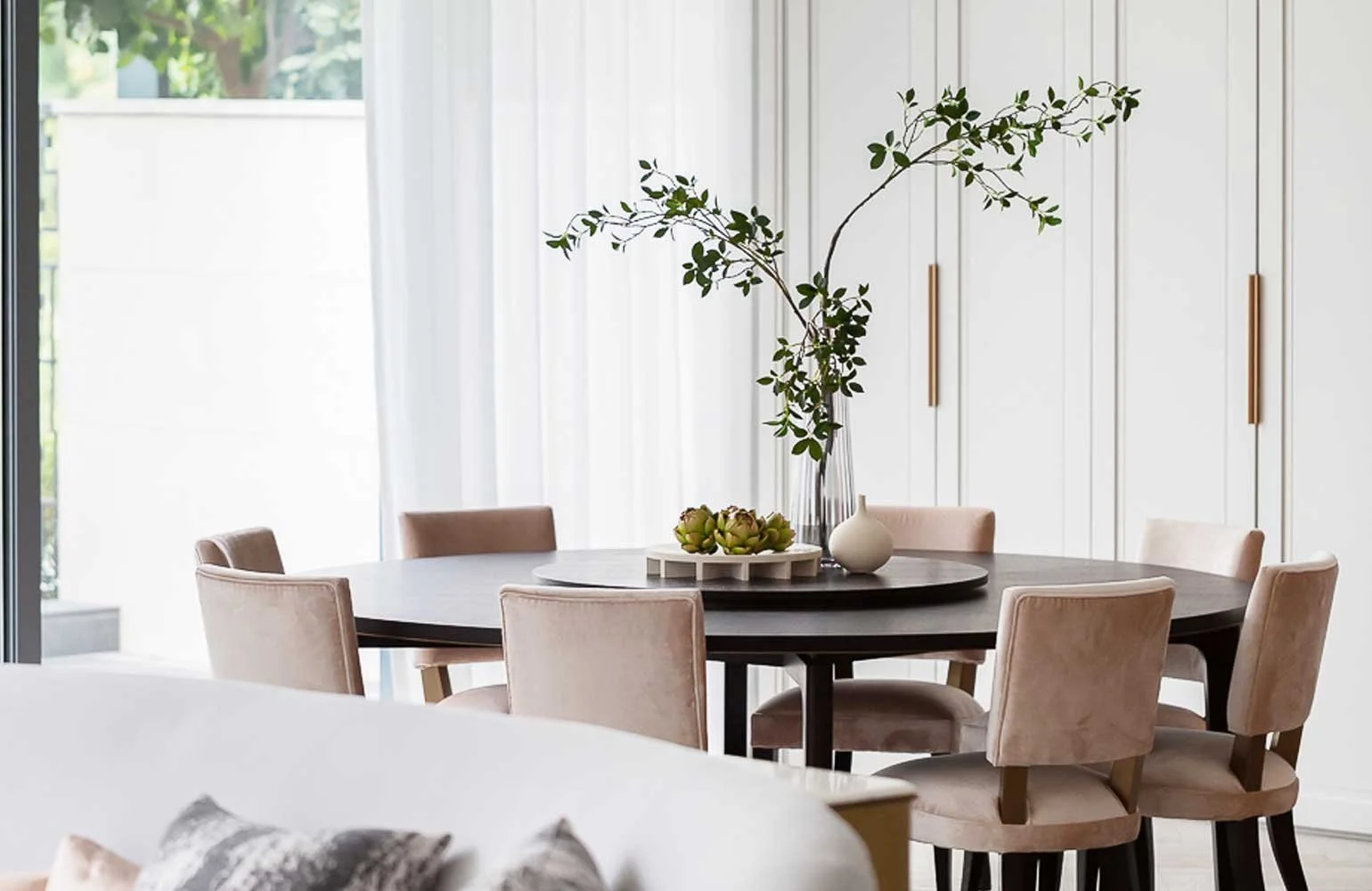 Round dining table with beige upholstered chairs, decorated with a tall glass vase with greenery, a white vase, and artichokes on a tray, in a bright room with white walls and large window.
