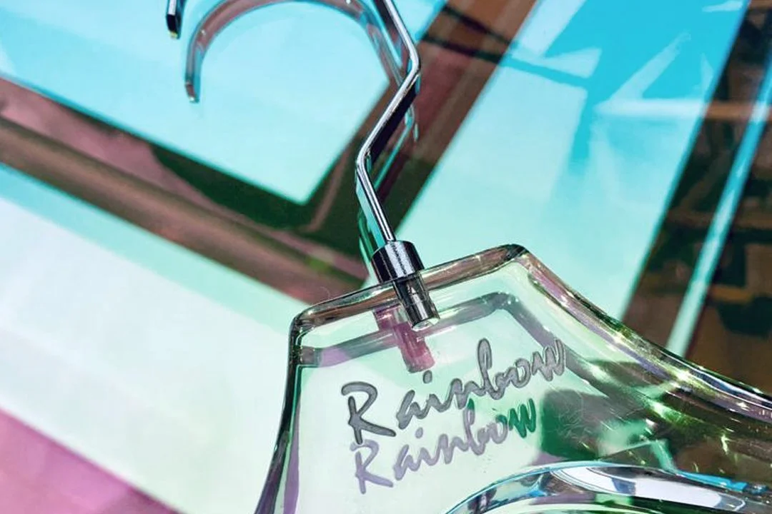 A close-up of a clear glass rainbow-shaped perfume bottle with silver accents, and the words 'Rainbow Rainbow' written on the glass in a cursive style, placed on a colorful pink and teal surface.
