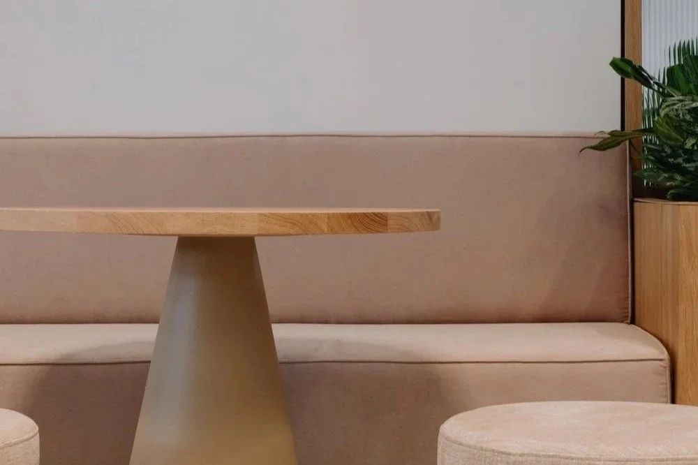 Minimalist interior with a beige upholstered bench, a round beige ottoman, a wooden table with a tapered base, and a potted plant in a wooden planter.