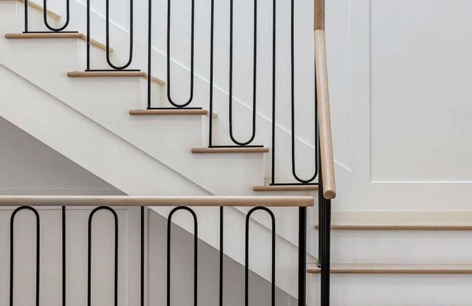 Interior view of a staircase with wooden steps, black metal balusters with U-shaped designs, and wooden handrails attached to the wall.