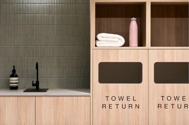 Laundry area with a sink, black faucet, soap dispenser, folded white towels, pink water bottle, and laundry bins labeled "Towel Return."