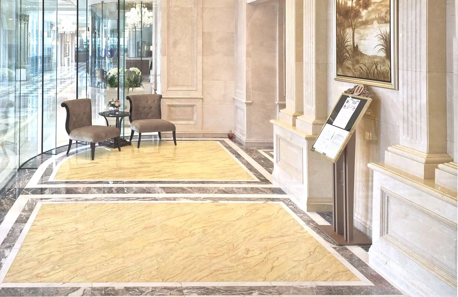 Elegant hotel lobby with marble flooring, two upholstered chairs, a small circular table with flowers, large window, framed artwork, and a signboard