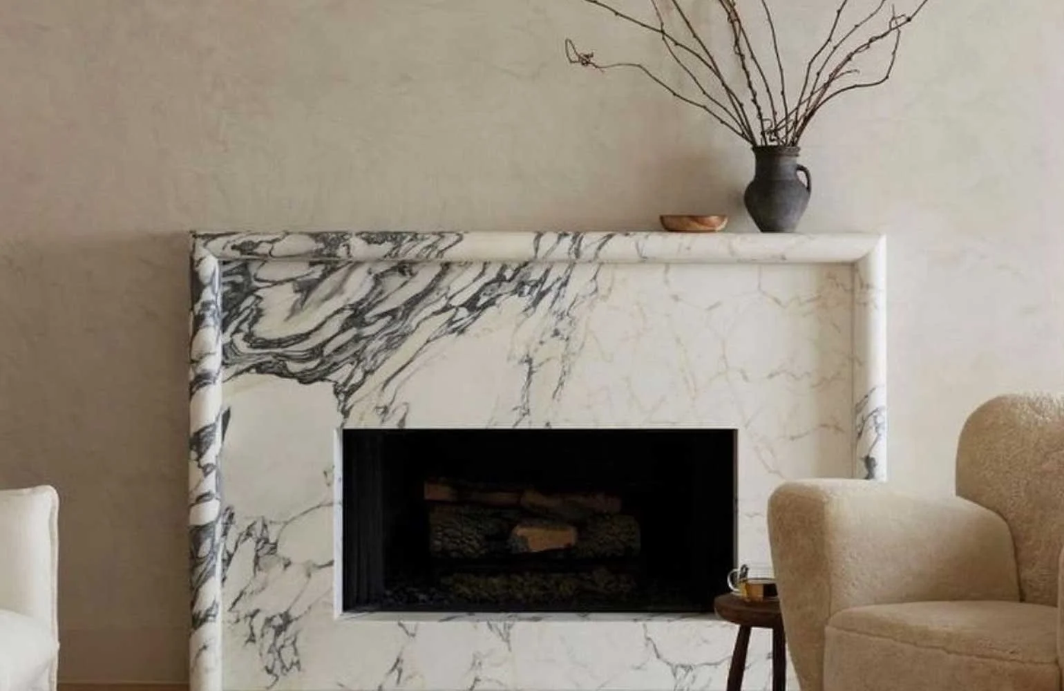 A white marble fireplace with black veining, topped with a black vase holding bare branches, a small wooden bowl, and surrounded by beige chairs.