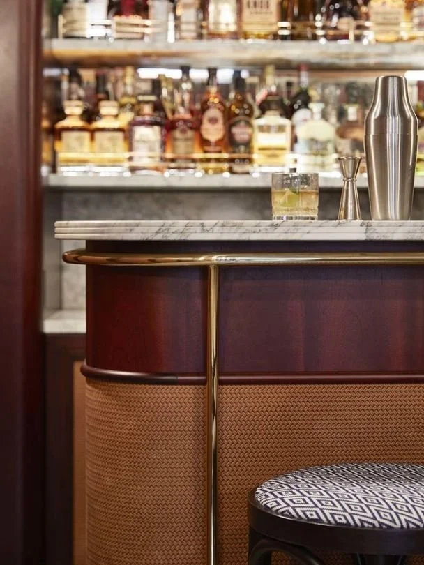 Bar counter with a marble top, a cocktail glass with a lime wedge, a stainless steel cocktail shaker, and a bartender's jigger, with shelves of liquor bottles in the background.