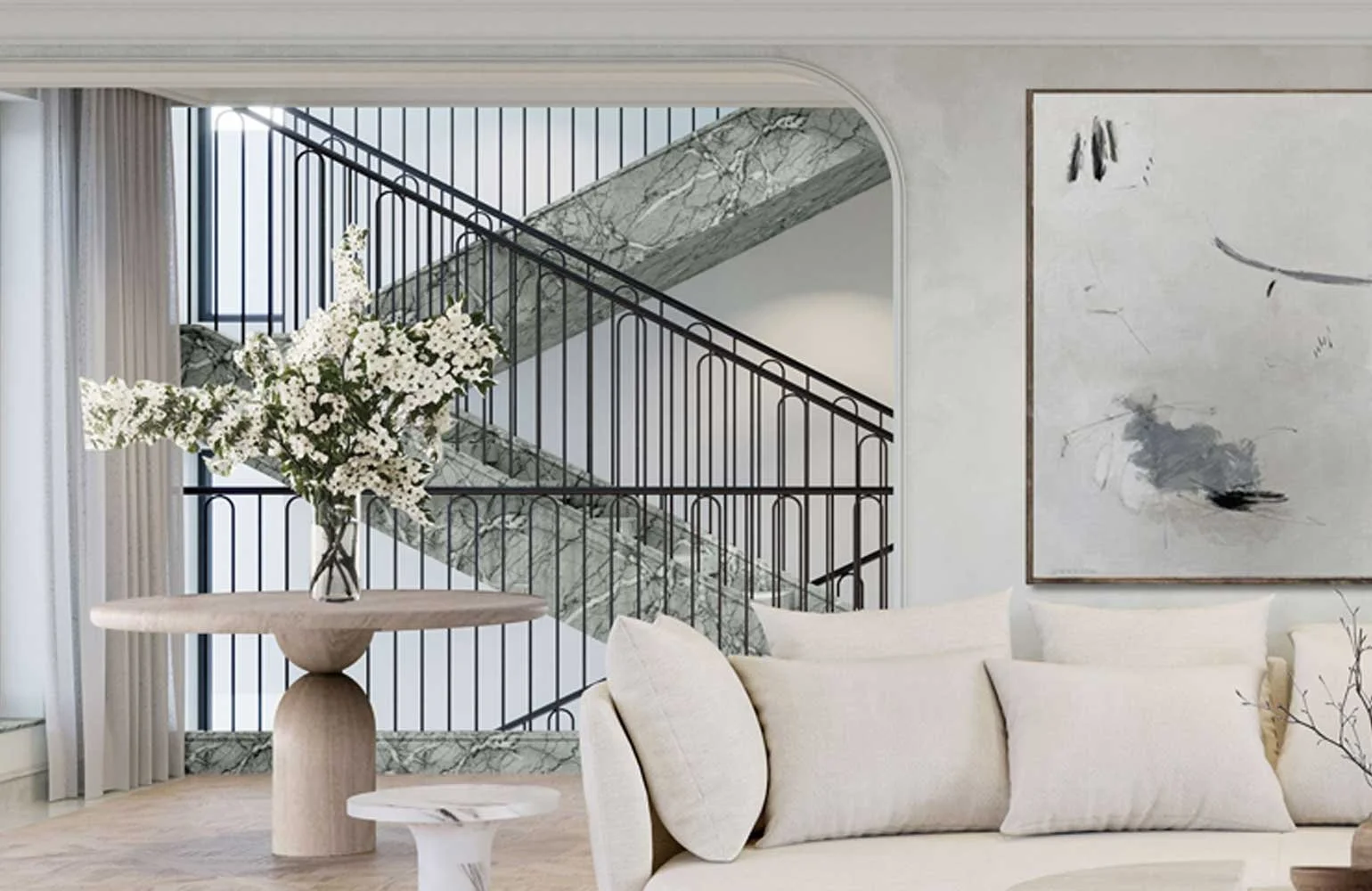Modern living room with a glass vase of white flowers on a round beige table, a cream sofa with pillows, a marble staircase with black railings, and abstract artwork on the wall.