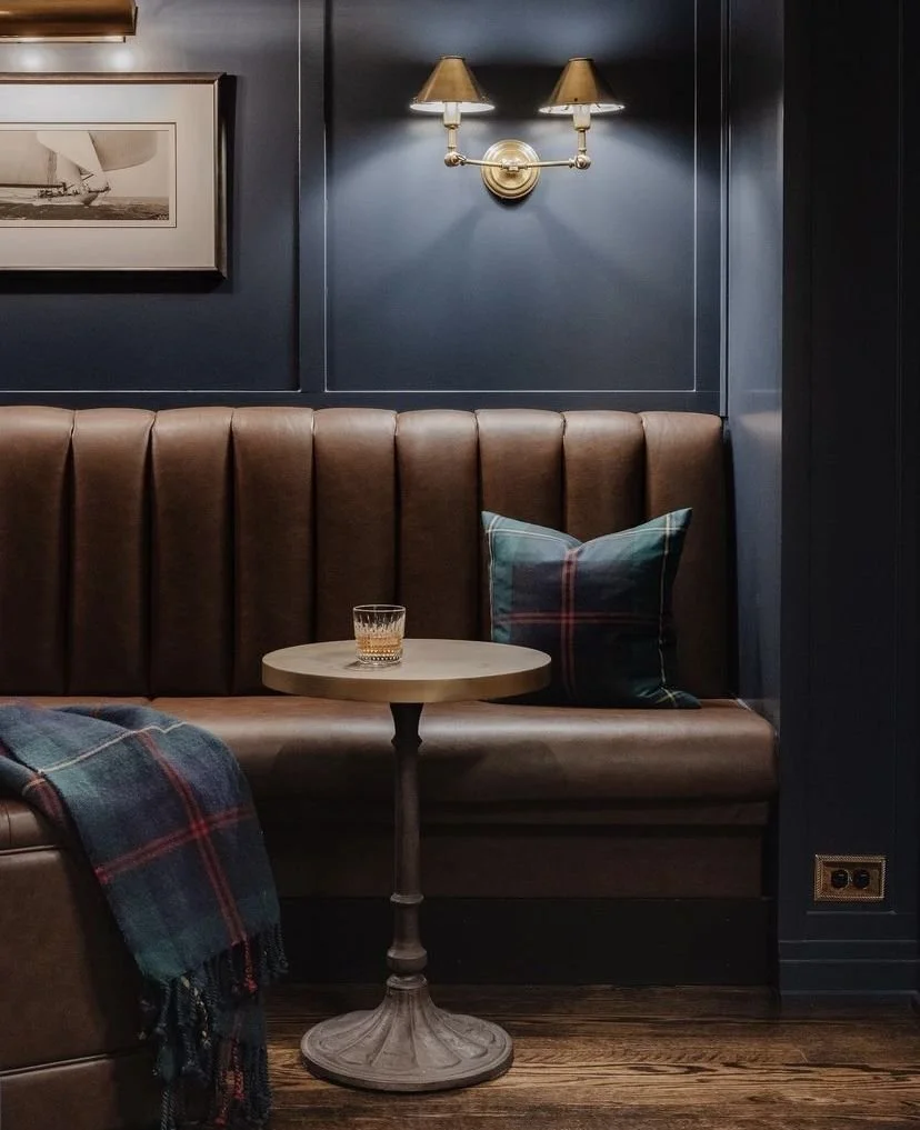 A cozy corner with a brown leather bench, dark blue wall, wall-mounted double lamps, a framed black-and-white photo, a small round wooden table with a glass of whiskey, a plaid cushion, and a plaid blanket.
