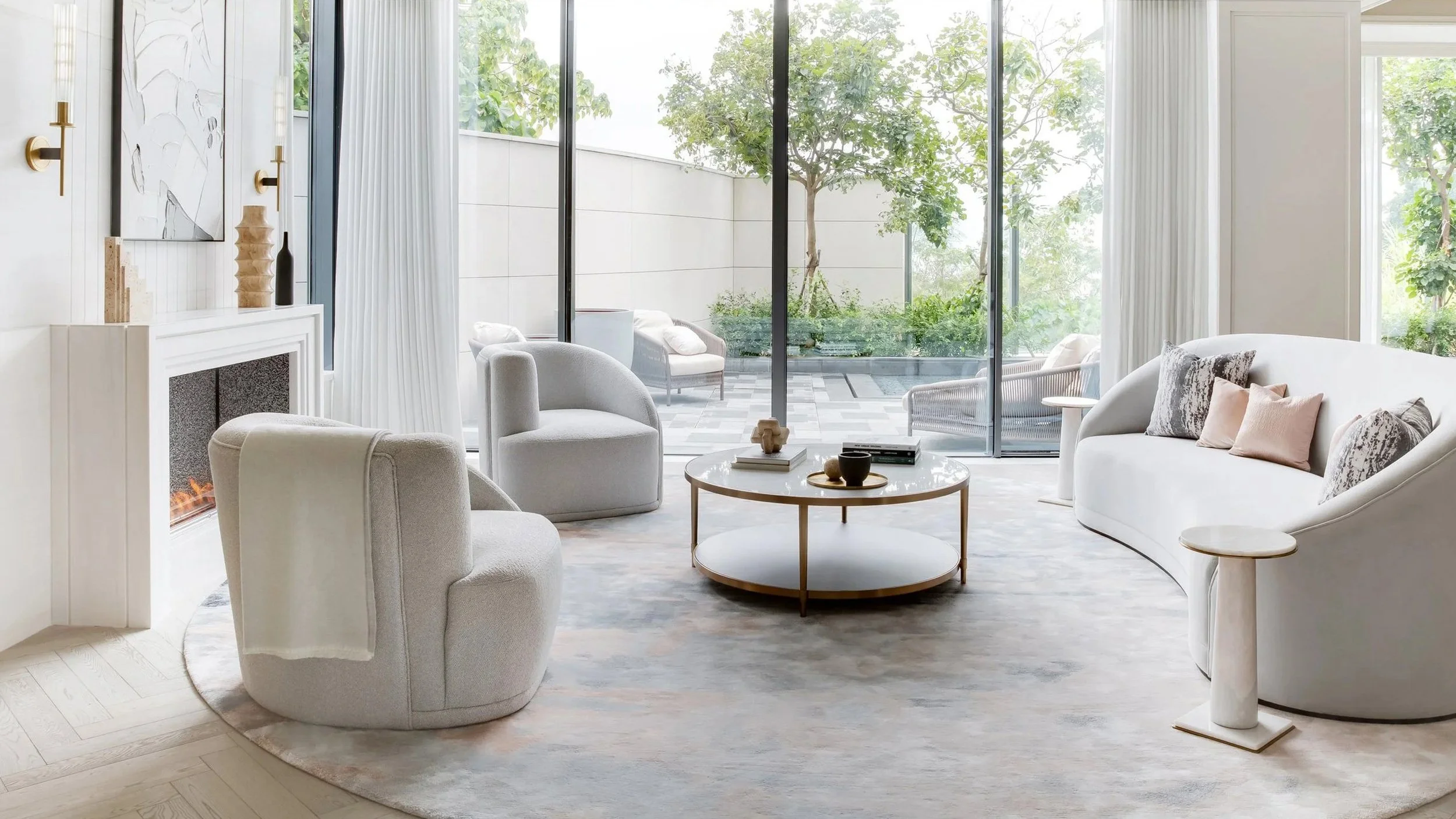 A modern living room with beige and white furniture, a fireplace, and large glass sliding doors leading to an outdoor patio with trees and greenery.