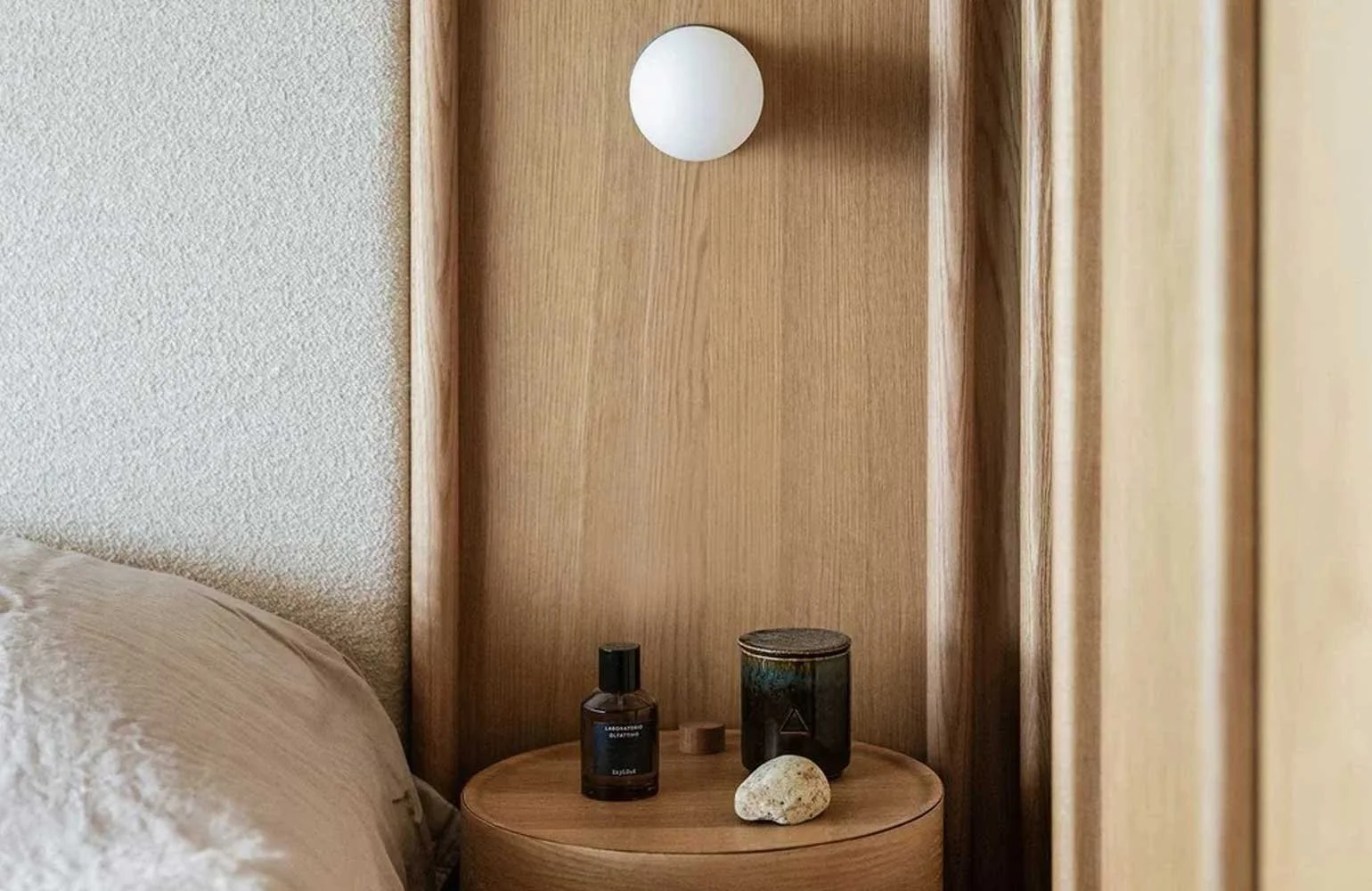 A round wooden bedside table with a black bottle, a black container, and a rock, positioned next to a bed with a beige sheet, against a wooden headboard and a textured wall.