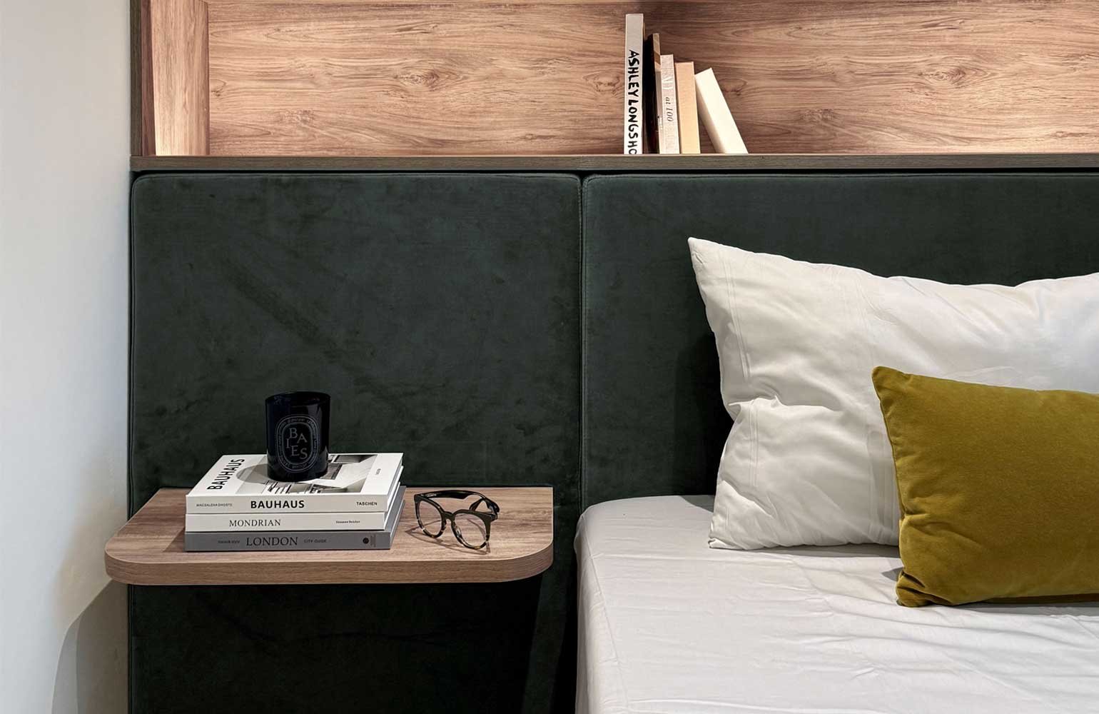 A bedside table with a book, a black candle, and glasses, next to a bed with white and yellow pillows, and a dark green headboard, in a modern bedroom with wooden accents.