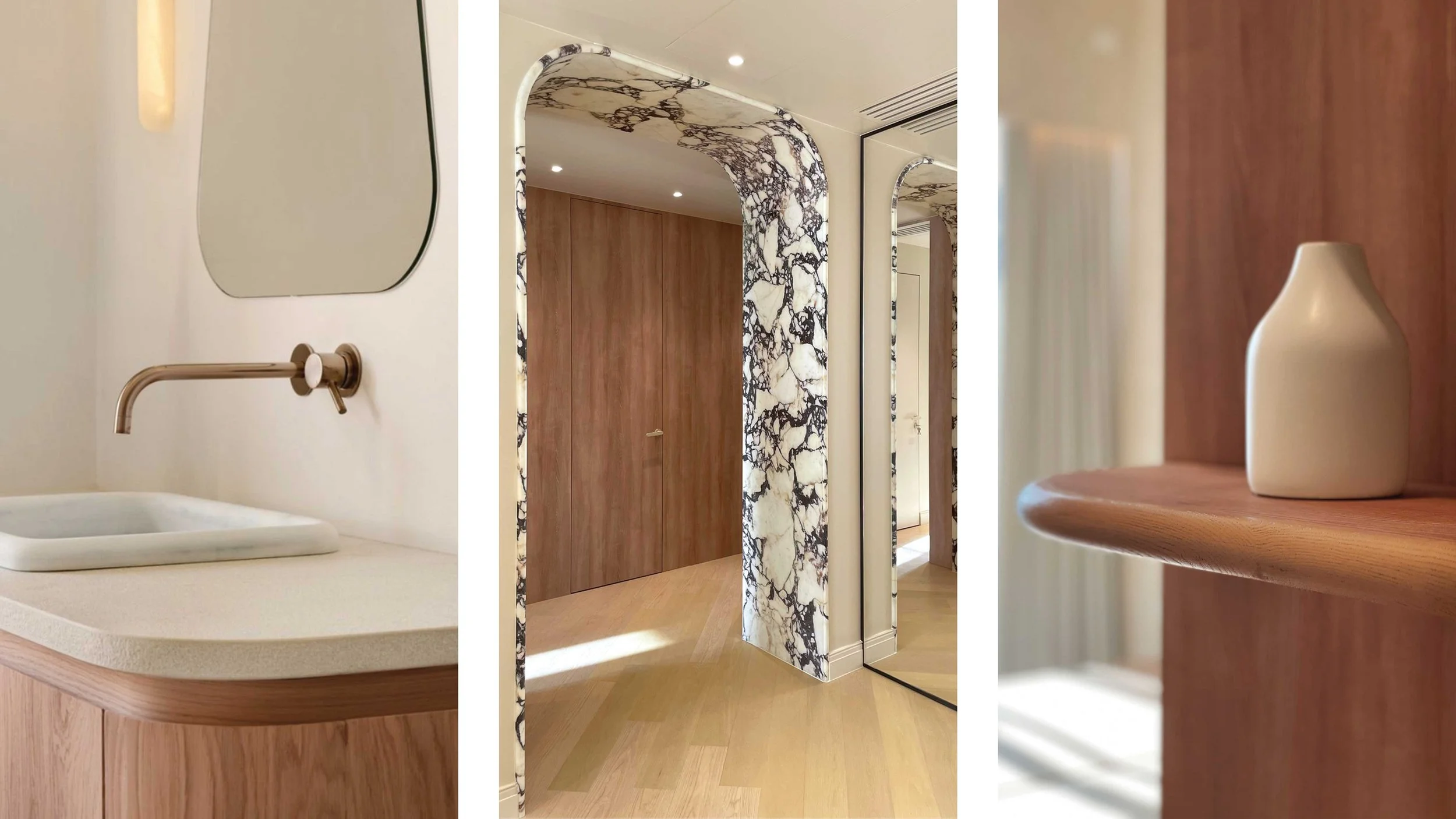 Close-up of a white bathroom sink with a modern brass faucet, a large irregularly shaped mirror, a wooden door, marble patterned architectural elements, and a wooden shelf holding a cream-colored vase.