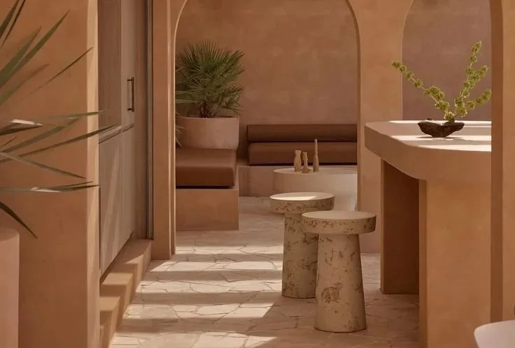 Interior of a stylish, minimalist lounge with tan walls, a built-in bench with cushions, potted plants, and small round tables with decorative items.