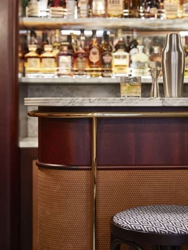 A bar counter with a marble top, a glass with a drink, and a metal cocktail shaker. Bottles on shelves in the background.